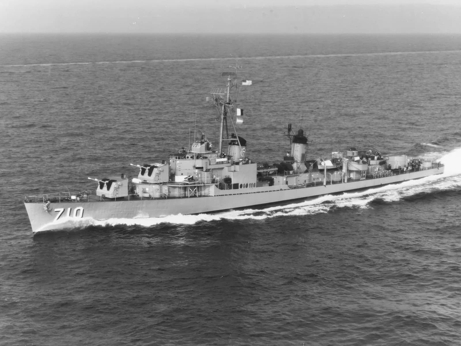 Aerial photograph of a grey destroyer with the hull number 710 cutting through a calm sea, its wake white behind it.