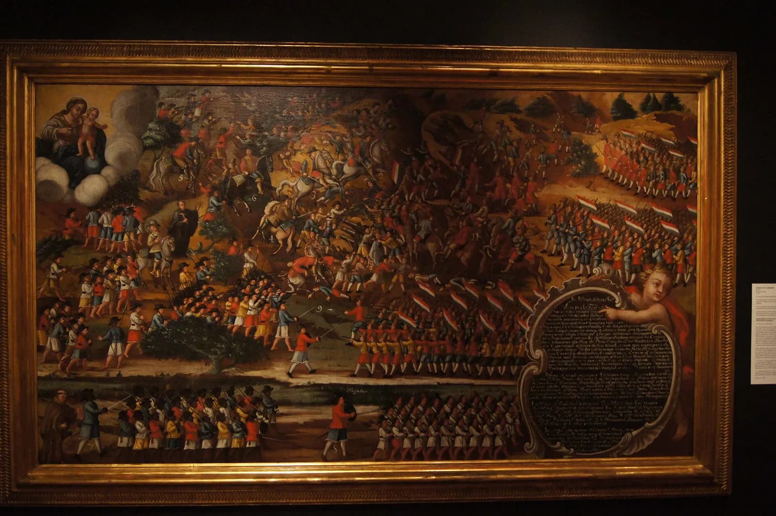 A framed painting crowded with small figures: ranks of soldiers under red, white and blue Dutch flags on the right face musketeers, horsemen and companies of Black soldiers on the left. The Virgin and Child sit on a cloud in the upper left corner, and a cherub holds a cartouche of text in the lower right.