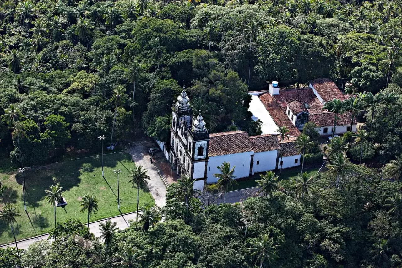 Aerial view of a white church with two ornate bell towers and a long tiled-roof convent building, standing beside a lawn with palm trees and surrounded by dense green woodland.