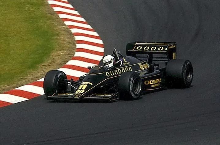 A black and gold Lotus Formula One car with a white-helmeted driver takes a corner beside red and white kerbs.
