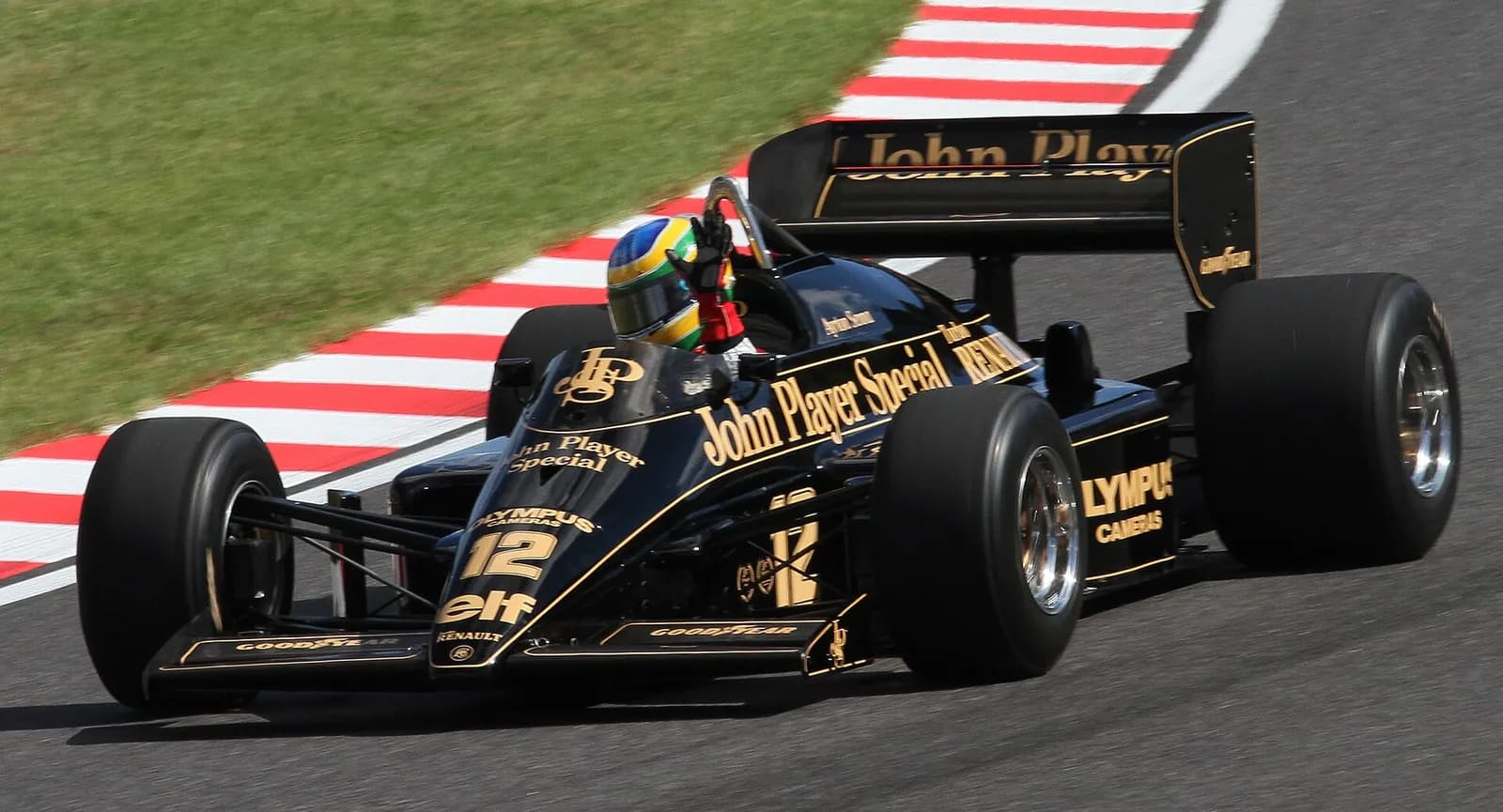 A black Lotus Formula One car with gold John Player Special lettering and the number 12 rounds a red and white kerb, its driver in a yellow, green and blue helmet raising a hand to wave.