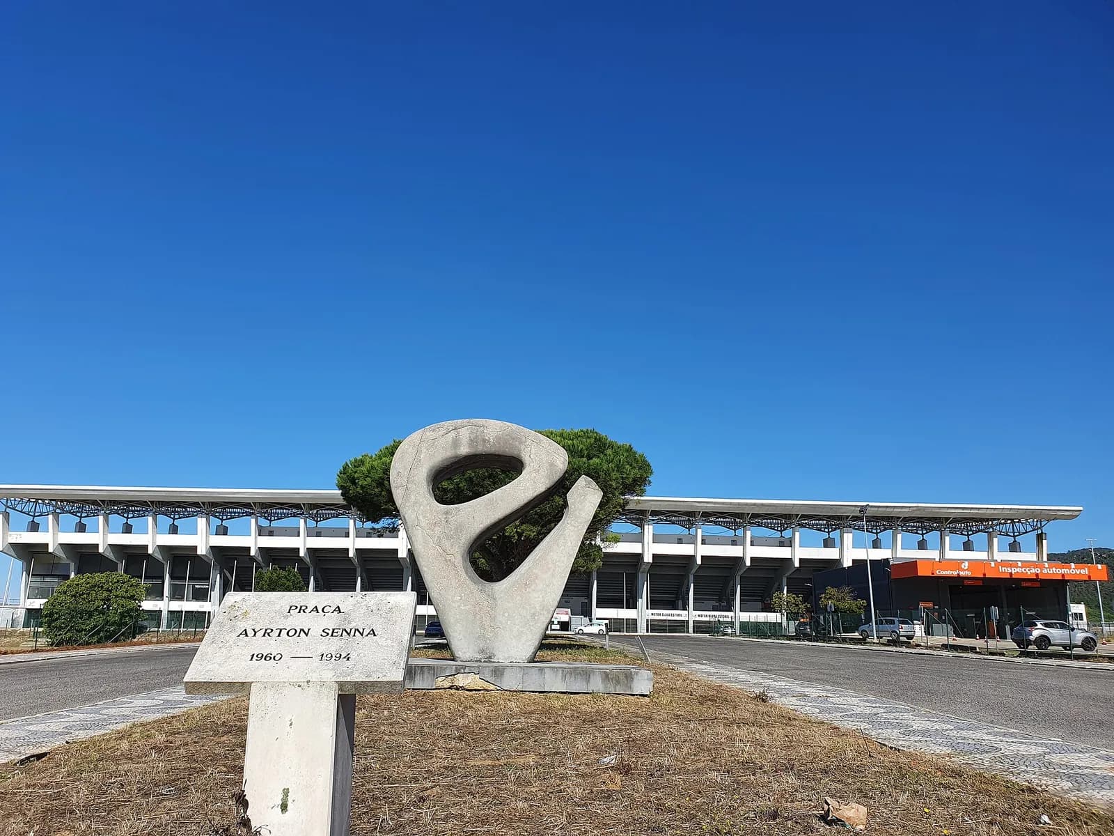 An abstract stone sculpture and a small stone marker reading Praça Ayrton Senna 1960–1994 stand in front of the long concrete main grandstand of the Estoril circuit under a clear blue sky.