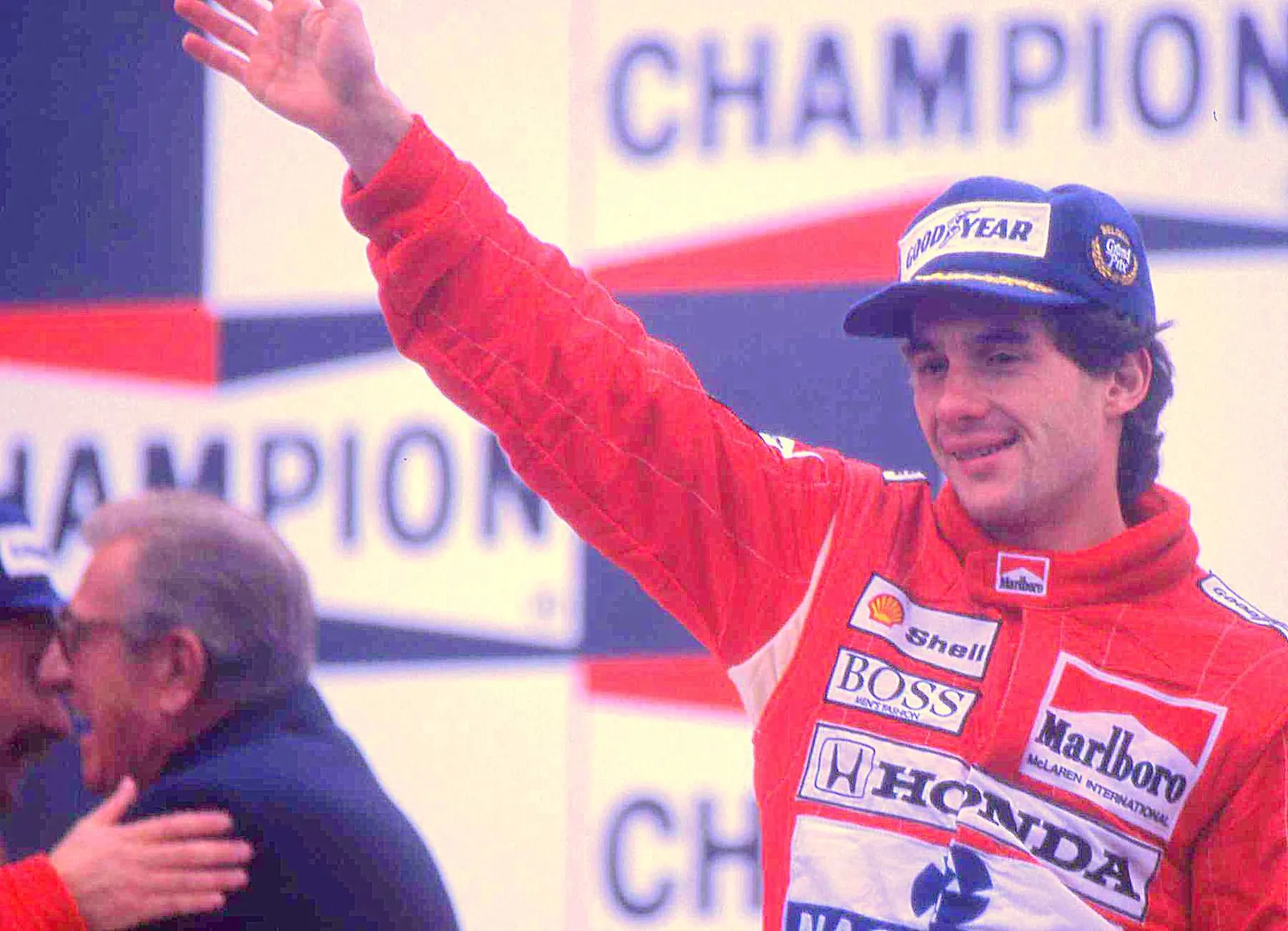 Ayrton Senna in red McLaren overalls and a blue Goodyear cap waves from a podium in front of a Champion banner.