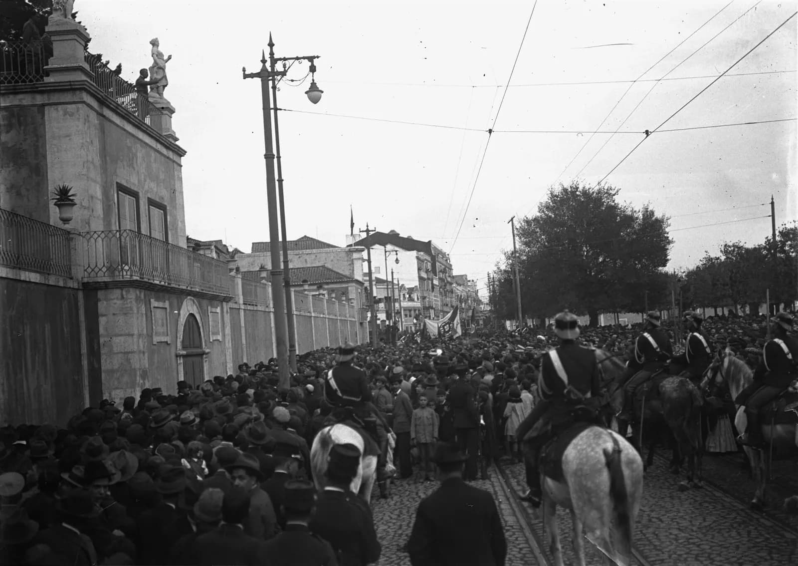 A dense crowd of men in hats fills a cobbled street beside a high palace wall topped by statues, watched by mounted guards in white cross-belts. Tram wires cross the sky and a flag rises above the crowd in the distance.