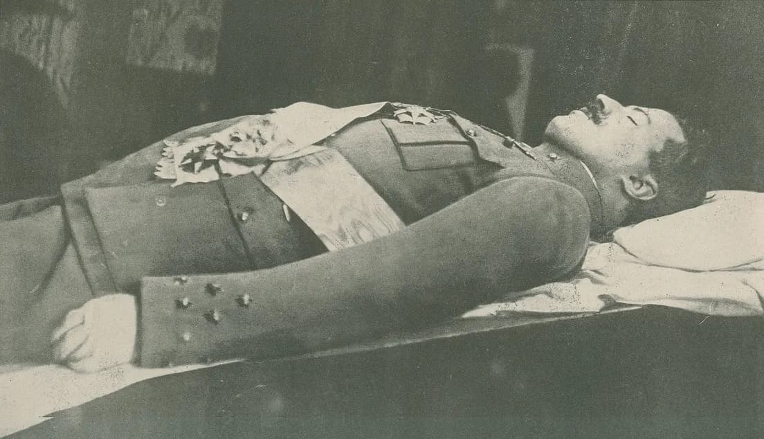 A dead man in a buttoned military tunic with stars on the cuffs lies on his back on a sheet-covered bier, his head on a white pillow and decorations on his chest.