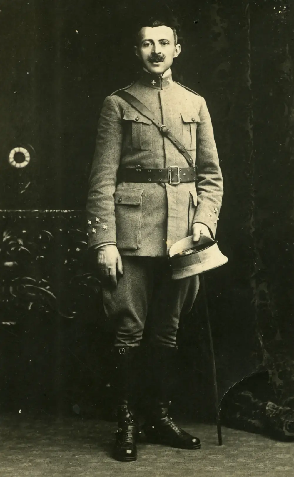 Full-length studio photograph of a slim man with a moustache in a belted field uniform with a shoulder strap and stars on the collar and cuffs, holding his cap in one hand, a thin cane at his side.