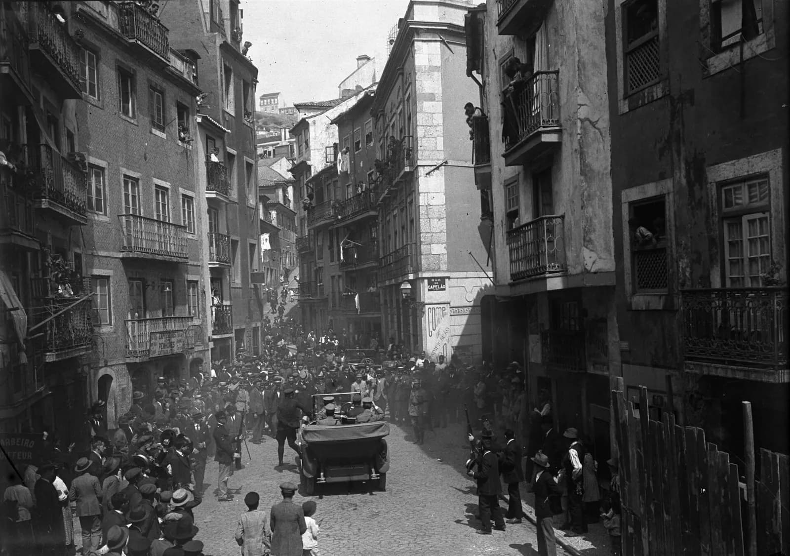 An open car moves slowly up a narrow cobbled street between tall old houses, surrounded by a dense crowd of men in hats and caps; people watch from balconies and windows.