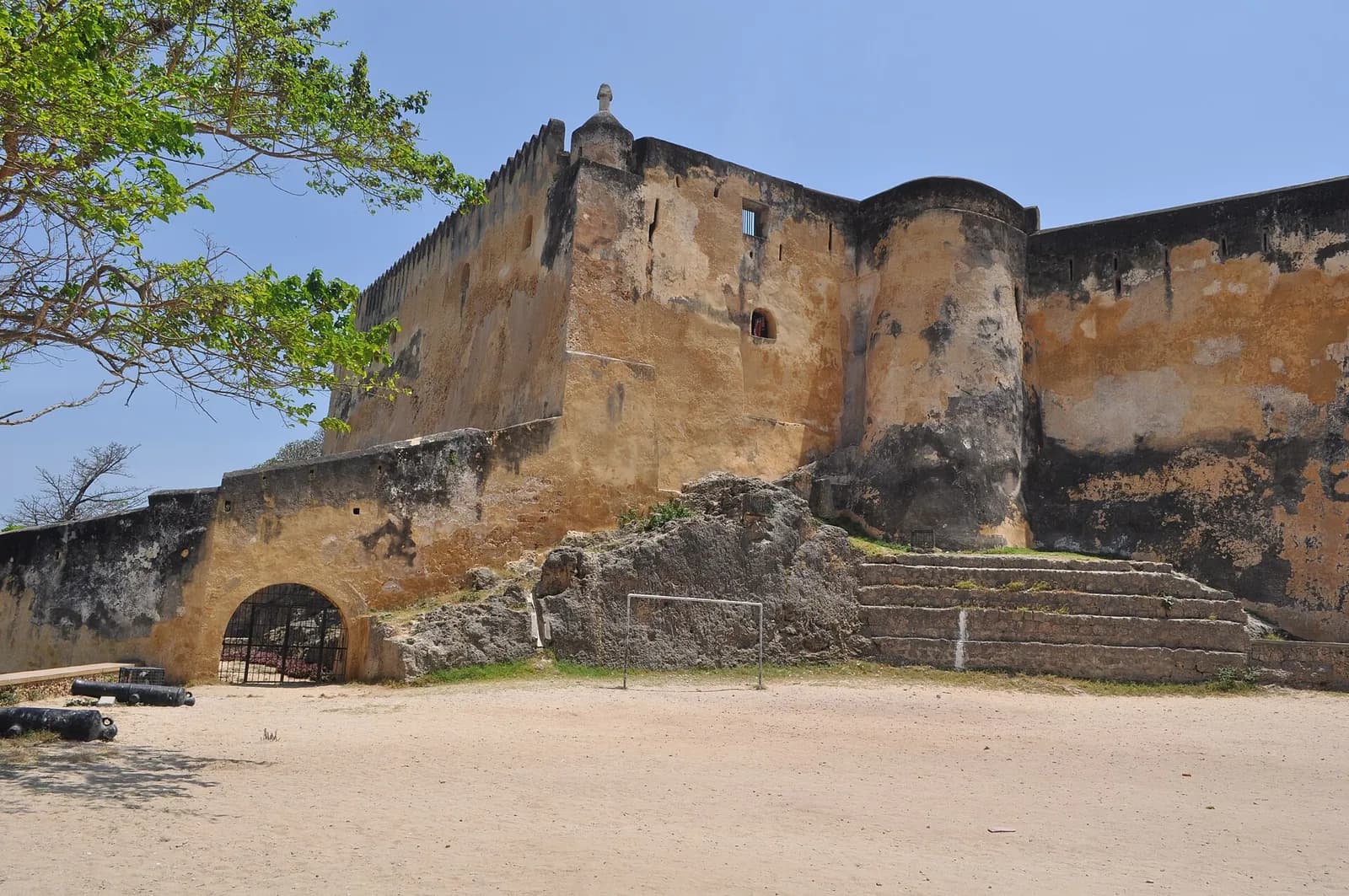 Tall, weathered ochre walls of a stone fortress under a blue sky, with a rounded tower in the middle, a small domed turret on the corner, a gated archway in a lower outer wall and a sandy open ground in front.