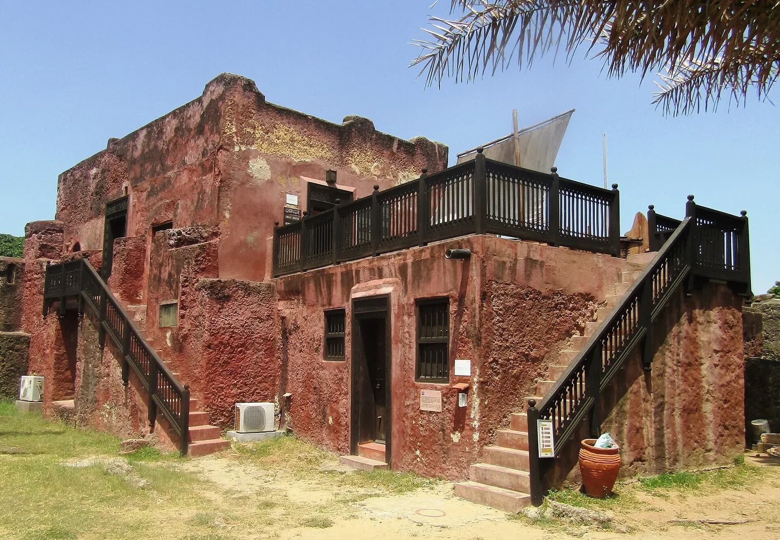 A two-storey building with weathered red-plastered walls inside a fort, with two outside staircases and a dark wooden balcony railing on the roof terrace, behind which the mast and sail of a dhow can be seen.