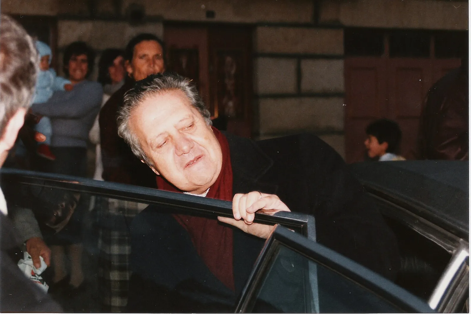 A grey-haired man in a dark coat and red scarf leans in at the open door of a dark car, one hand on the window frame. Behind him, smiling women, one holding a baby in a blue knitted suit, and a small boy stand in front of a stone house.