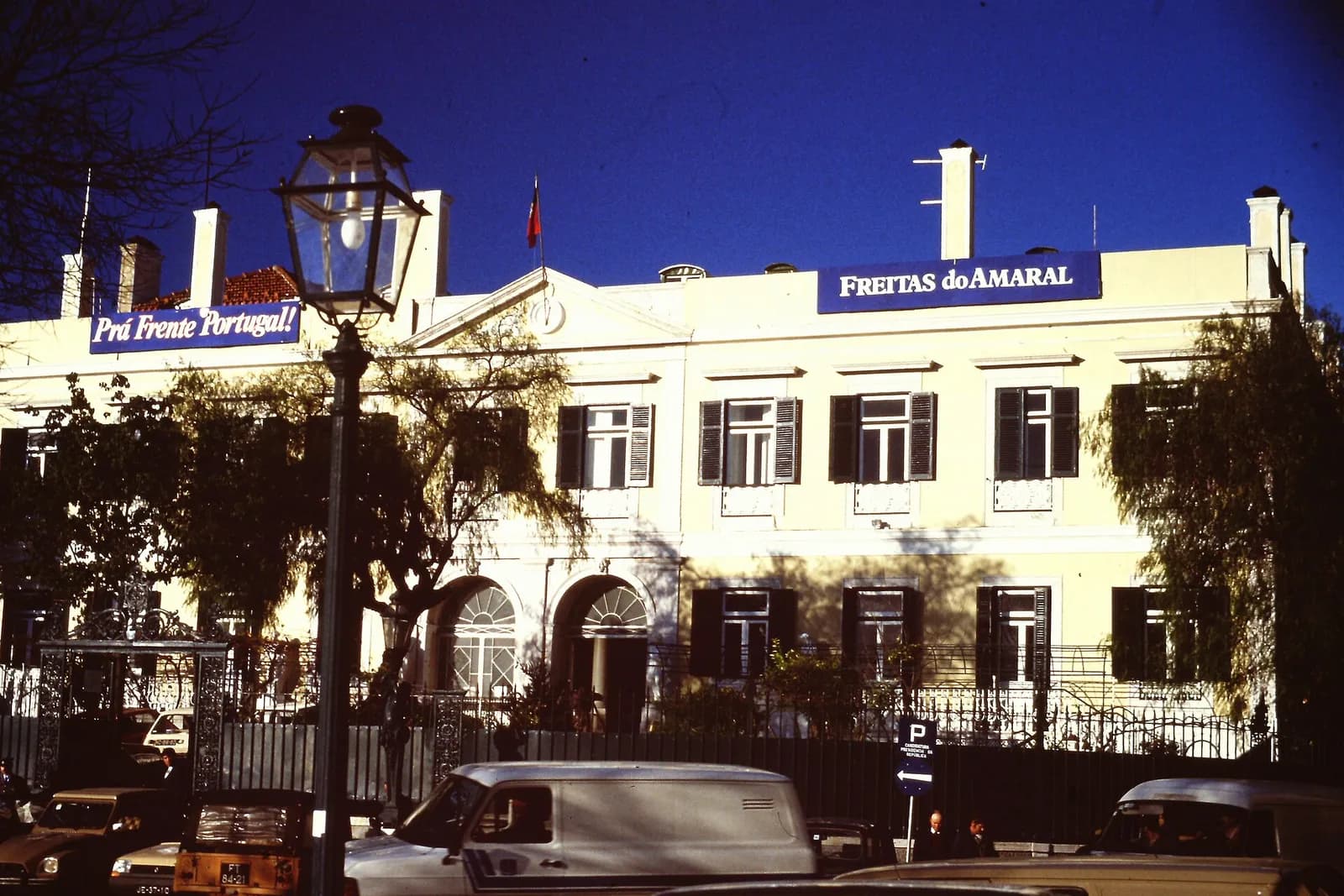 A pale two-storey building with green shutters behind railings and parked cars, under a deep blue sky. Blue banners along the roofline read Prá Frente Portugal! and Freitas do Amaral.