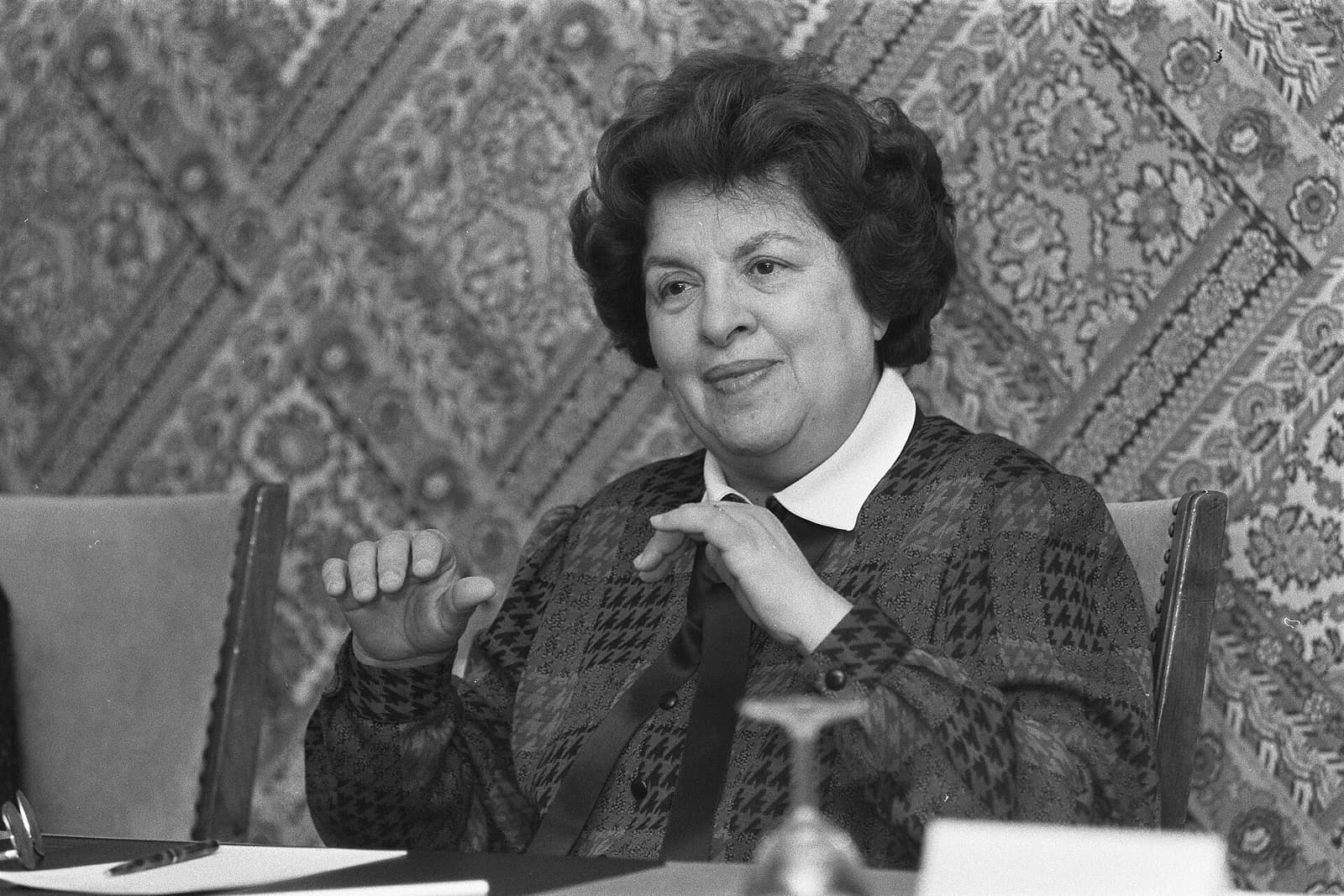 A woman with short dark curly hair, wearing a patterned blouse with a white collar, sits at a table gesturing with both hands in front of a patterned wall hanging.