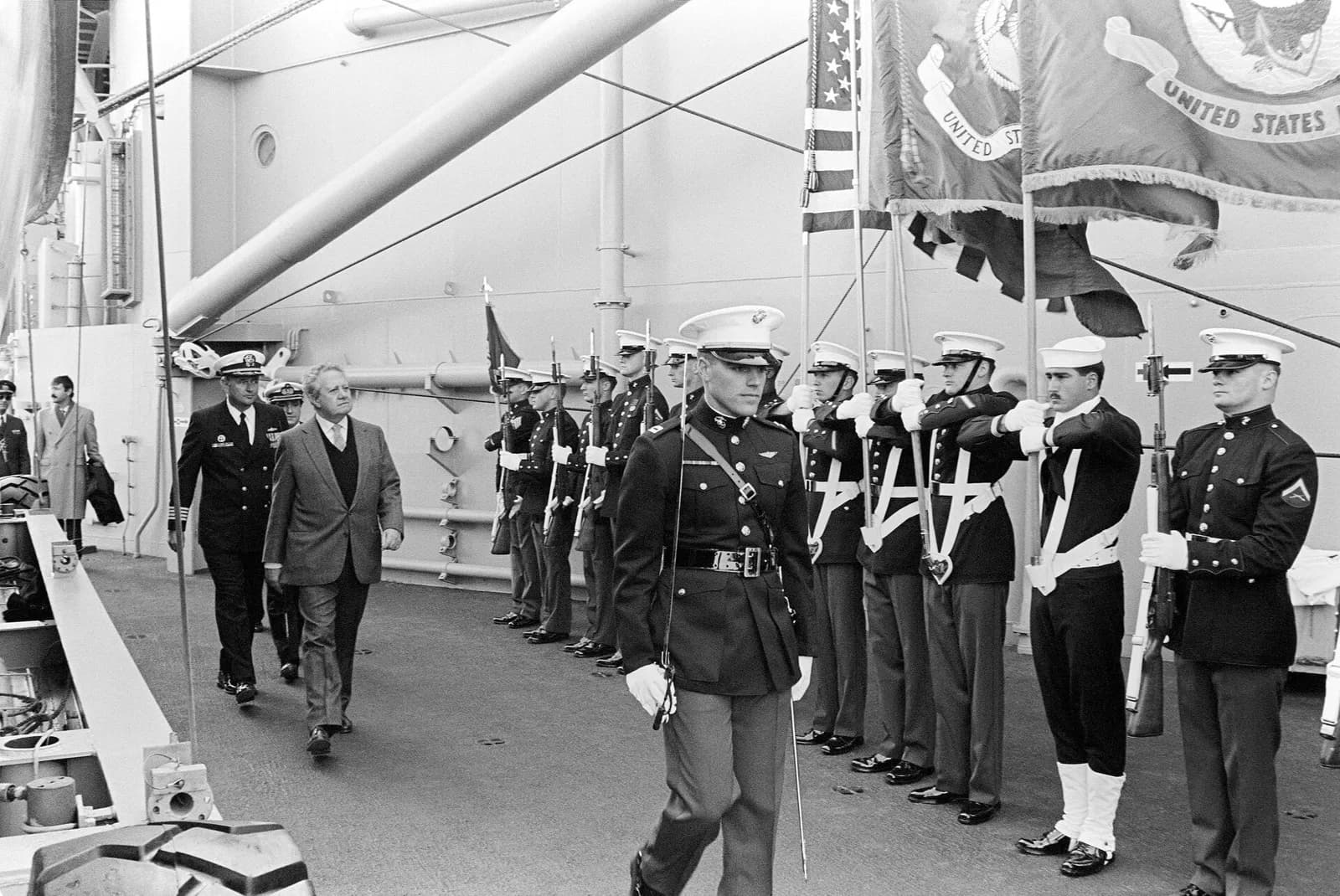 A grey-haired man in a jacket and tie walks beside a naval officer along the deck of a warship, past a line of American marines and sailors in dress uniform holding rifles and flags.