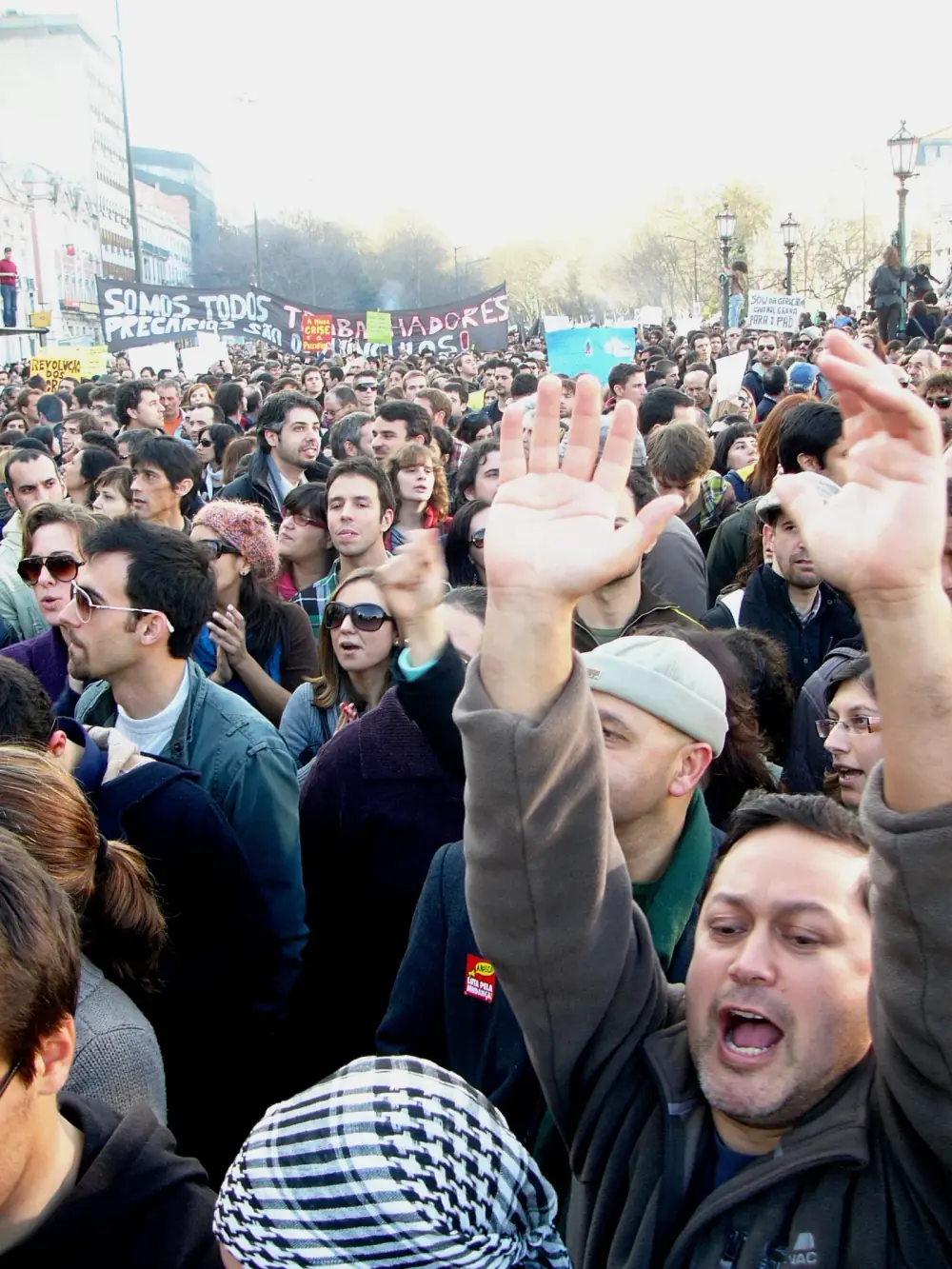 A packed crowd fills a wide tree-lined avenue under a hazy sky. In the foreground a man shouts with his arm raised, and banners are held up further back.