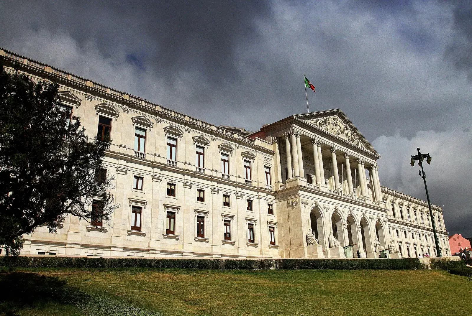 The long white neoclassical front of a large palace with a columned portico and a triangular pediment, under dark clouds. A green and red flag flies above the portico and a lawn slopes down in the foreground.