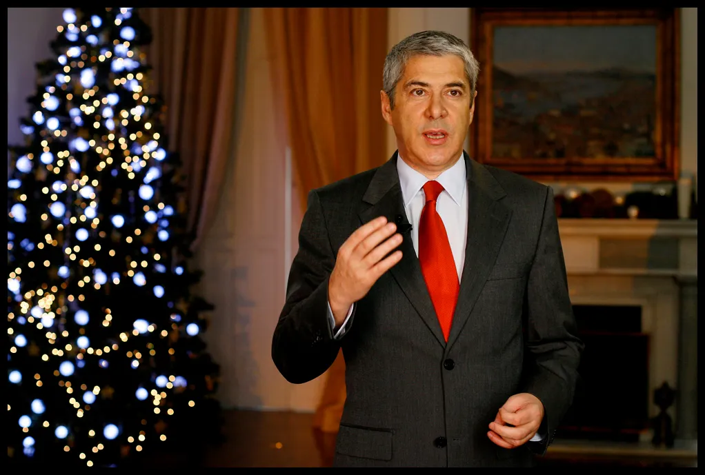 A grey-haired man in a dark suit and red tie speaks to camera with one hand raised, beside a Christmas tree hung with white lights, in a room with curtains and a framed painting.