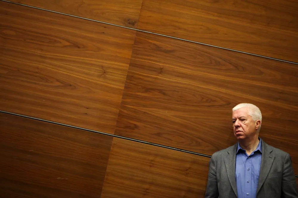 A white-haired man in a grey jacket and open-necked blue shirt stands at the lower right of the frame, looking to one side, against a wall of dark wood panels.