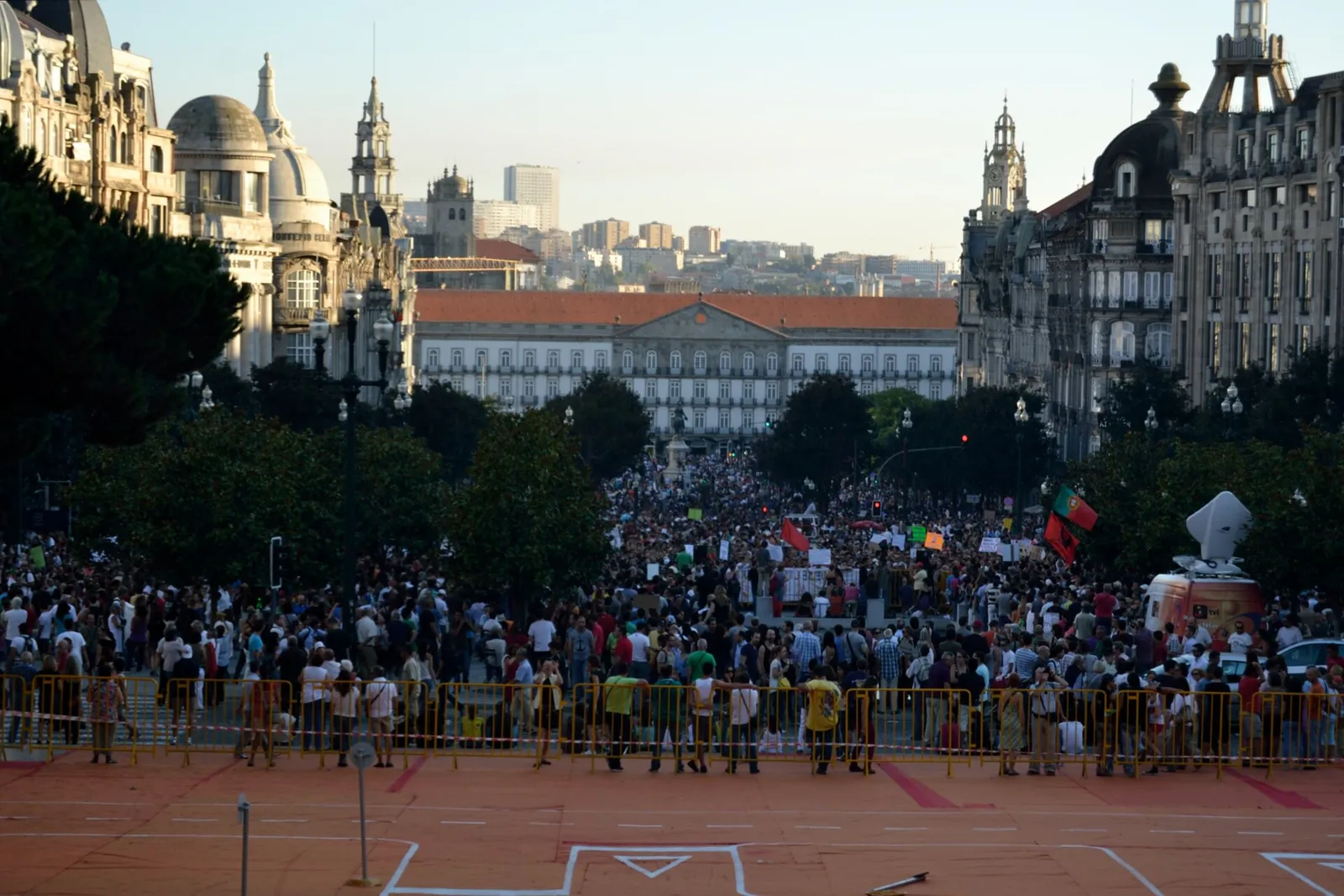 Seen from above, a large crowd fills a long avenue lined with grand stone buildings and trees, with a row of yellow barriers across the foreground and a long, low palace-like building at the far end.