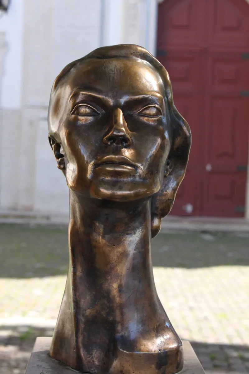 A bronze bust of a young woman with wavy hair swept back, a long neck and calm, half-closed eyes, standing outdoors in front of a white wall and a red door.