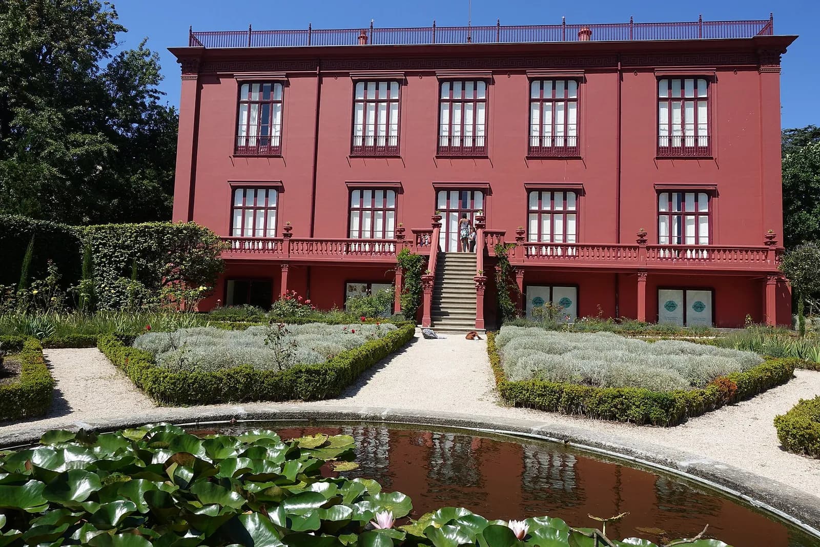 A tall three-storey house painted dark red, with a balustraded terrace and a central flight of steps, behind a formal garden of low clipped hedges, with a round pond and water-lily leaves in the foreground.