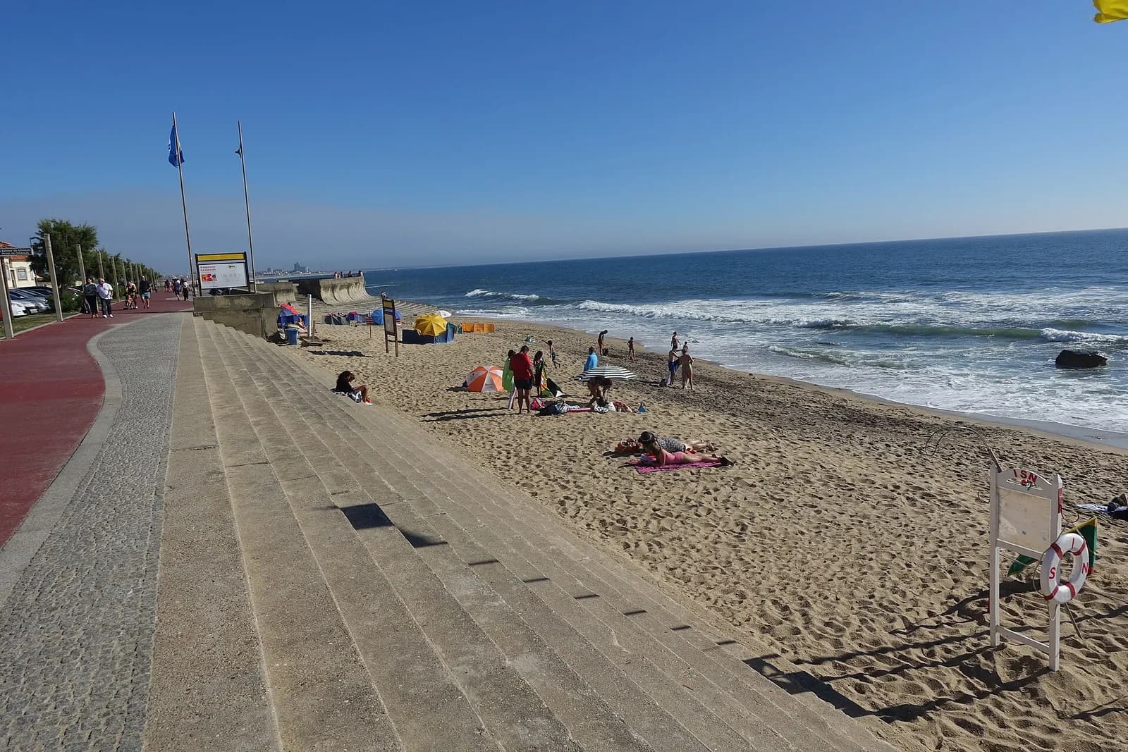 A wide sandy beach under a clear blue sky, with low waves breaking on the right, bathers and parasols on the sand, and broad stone steps and a promenade running along the left.