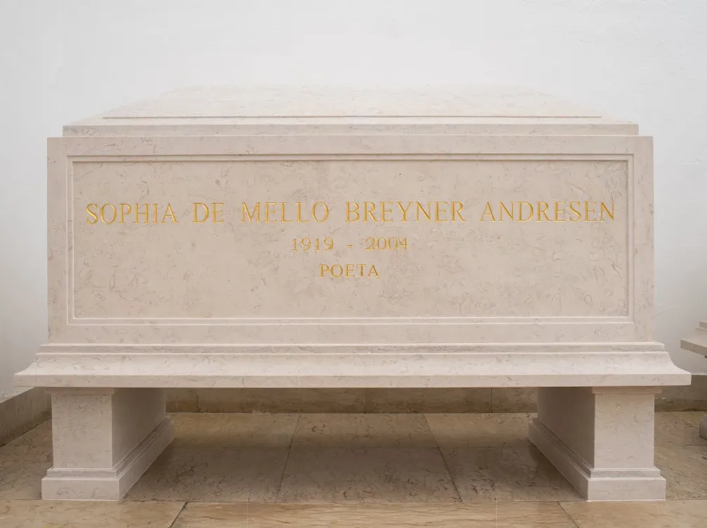 A plain cream limestone tomb chest on two square feet against a white wall, carved in gold capitals with the name Sophia de Mello Breyner Andresen, the dates 1919 to 2004 and the word Poeta.