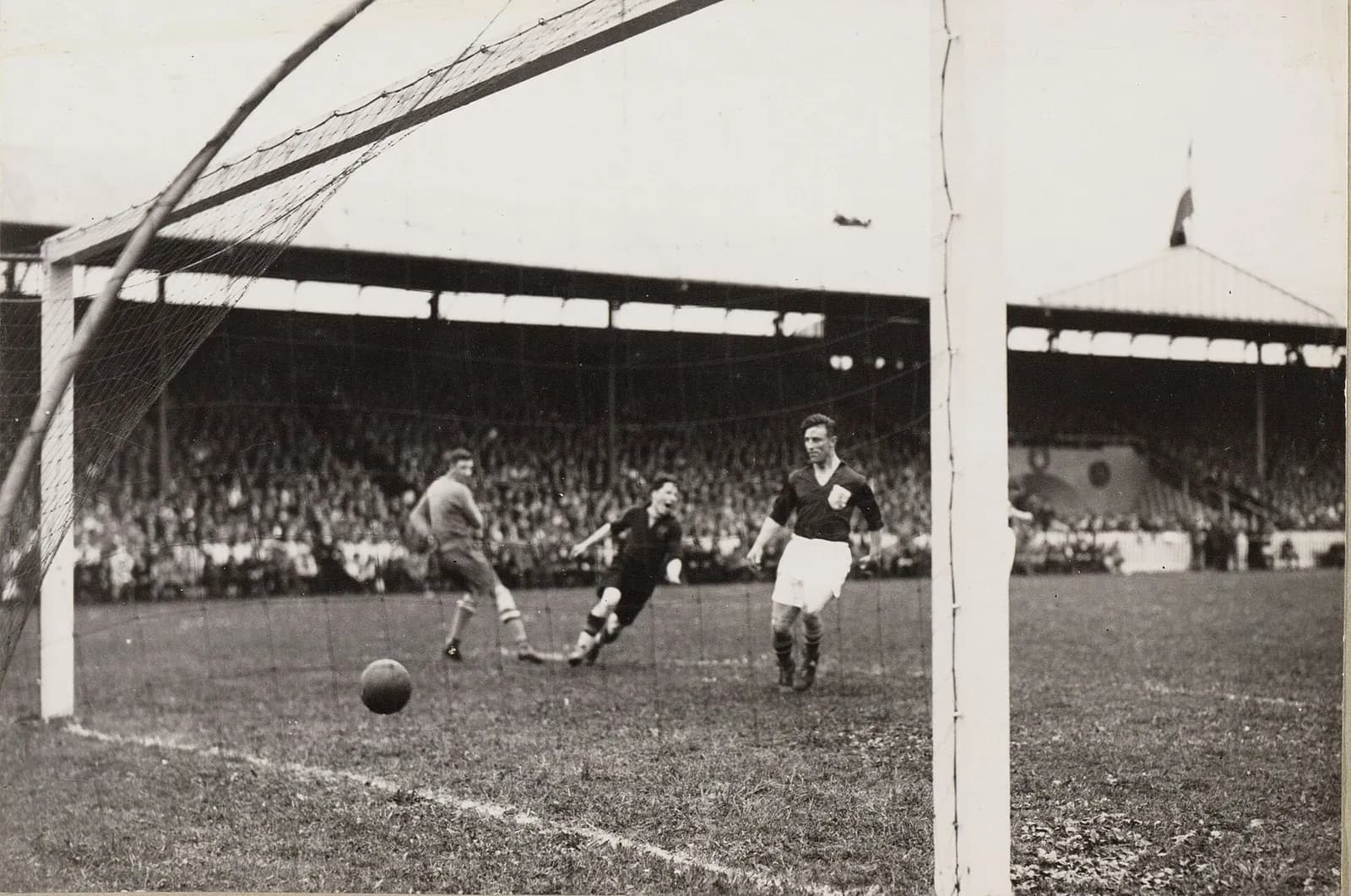 A black-and-white photograph taken from behind a goal net: the ball rolls towards the goal line as a goalkeeper in a light jersey turns away and two players in dark shirts follow it in, with a long covered stand full of spectators behind them.
