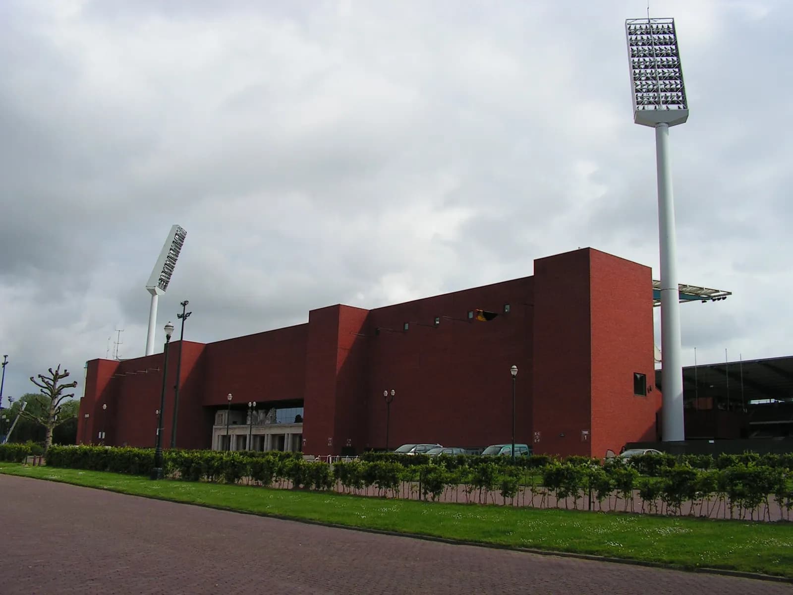 The red-brick outer wall of a stadium with two tall floodlight masts, seen across a lawn and a hedge under a grey sky.