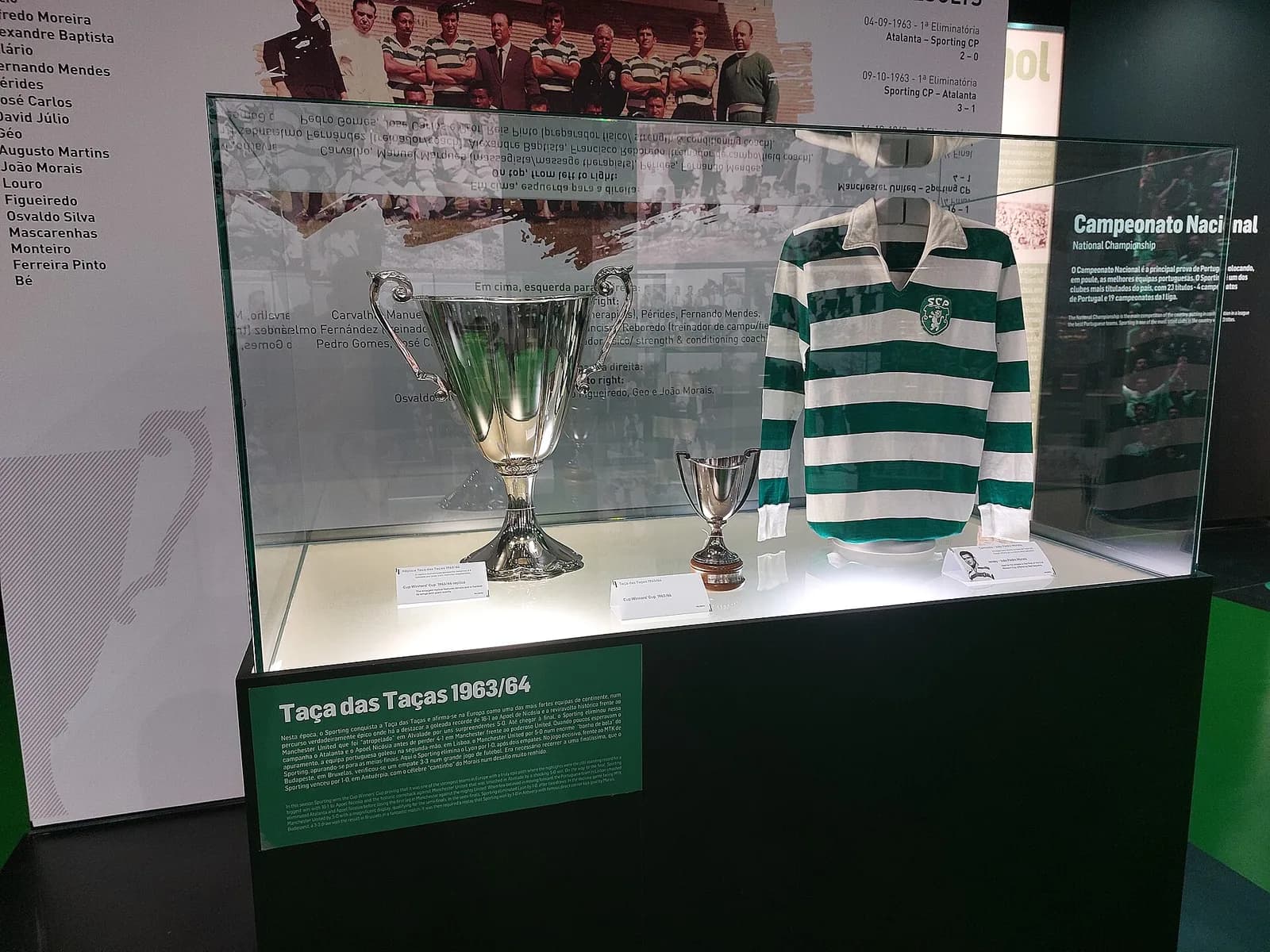 A lit glass case holds a large silver cup with curled handles, a much smaller silver cup on a wooden base and a long-sleeved shirt with green and white hoops. Behind it, a printed wall shows a black-and-white team photograph and a list of player names.