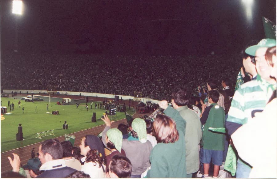 A packed stadium at night, seen from the terraces: supporters in green and white hoops and green hats cheer in the foreground, floodlights shine over a pitch ringed by a running track with vehicles and equipment on the grass, and a dark mass of spectators fills the far stand.