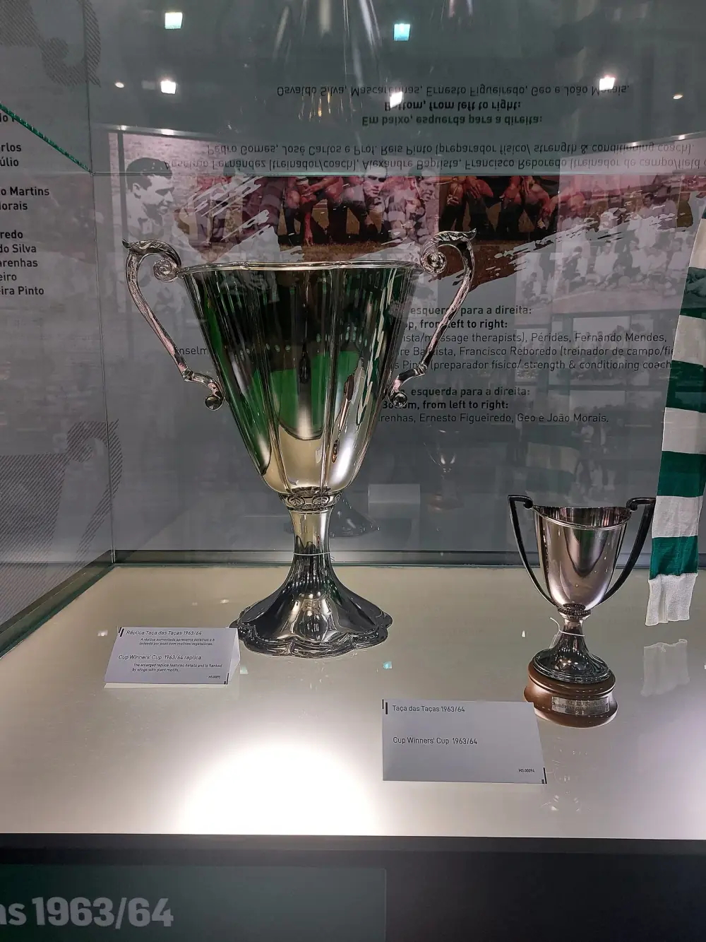 Inside a glass case, a large polished silver cup with tall curled handles stands on the left and a much smaller two-handled silver cup on a wooden base stands on the right, each with a small label. A green and white striped scarf hangs at the edge.