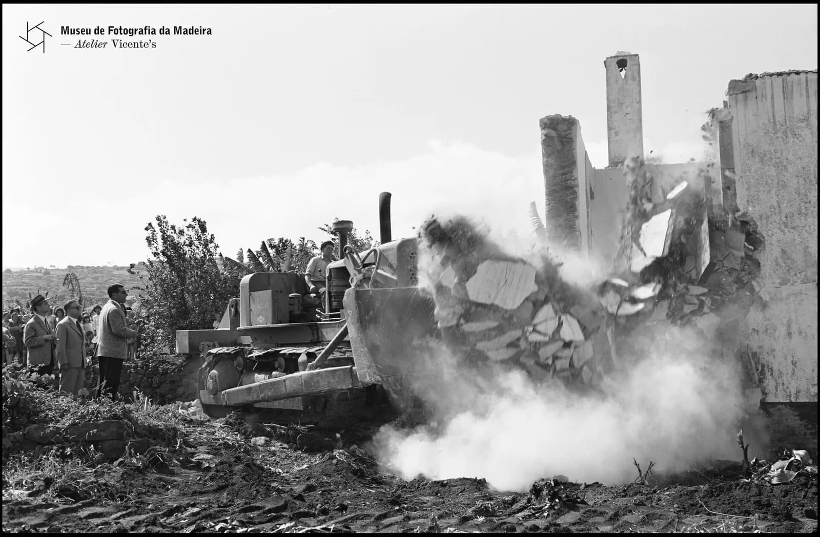 Black and white photograph of a crawler bulldozer pushing down the stone wall of a house in a cloud of dust and rubble. Men in jackets and a crowd of onlookers watch from the left, with terraced fields behind.