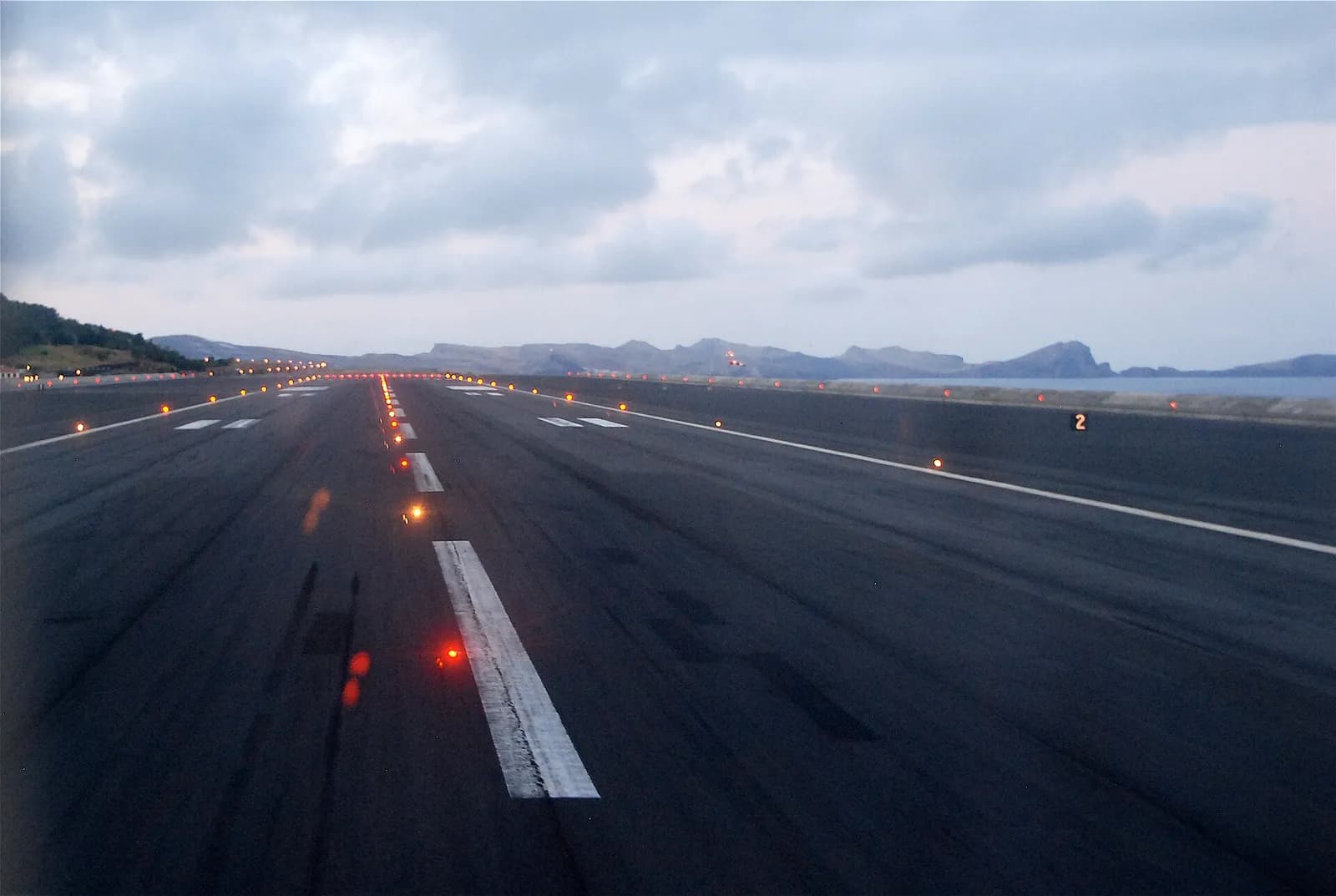 View along a dark asphalt runway at dusk, with white centreline markings and rows of glowing edge and centre lights leading towards low hills and the sea under heavy grey cloud.