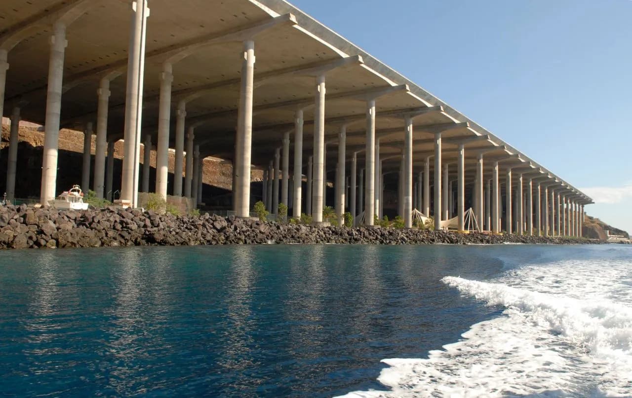 Seen from a boat, a flat concrete deck supported by long rows of slender white pillars runs along a rocky shore, with deep blue water and a boat's white wake in the foreground.