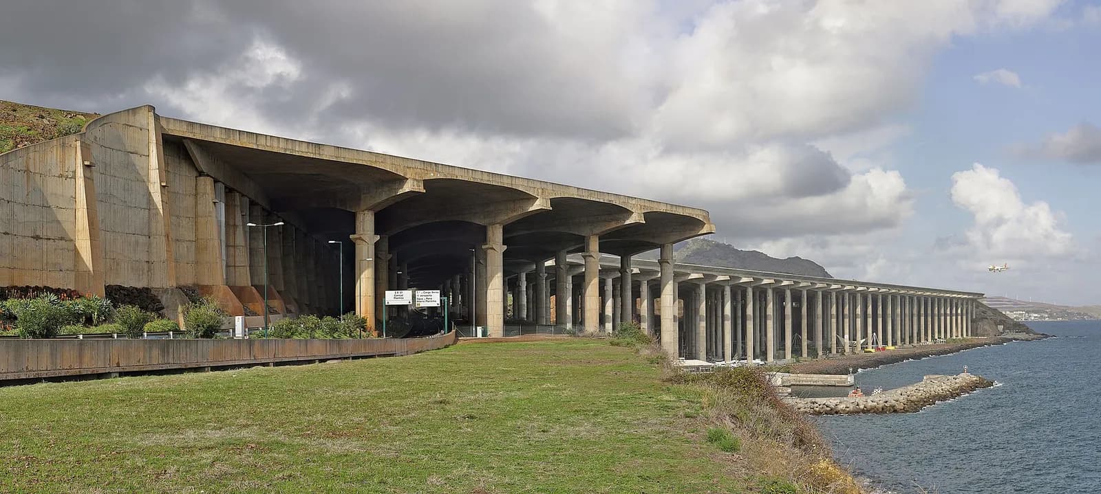 A long concrete deck held up by rows of tall pillars runs along a rocky shoreline beside the sea. A road passes beneath it, a grassy bank is in the foreground and an airliner is coming in to land on the right under a cloudy sky.