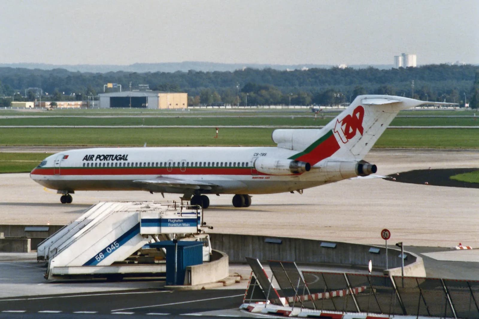A white airliner with a red and green stripe along the windows and three engines grouped at the tail taxis on an airport apron. The tail carries the red TAP logo and the fuselage reads Air Portugal.