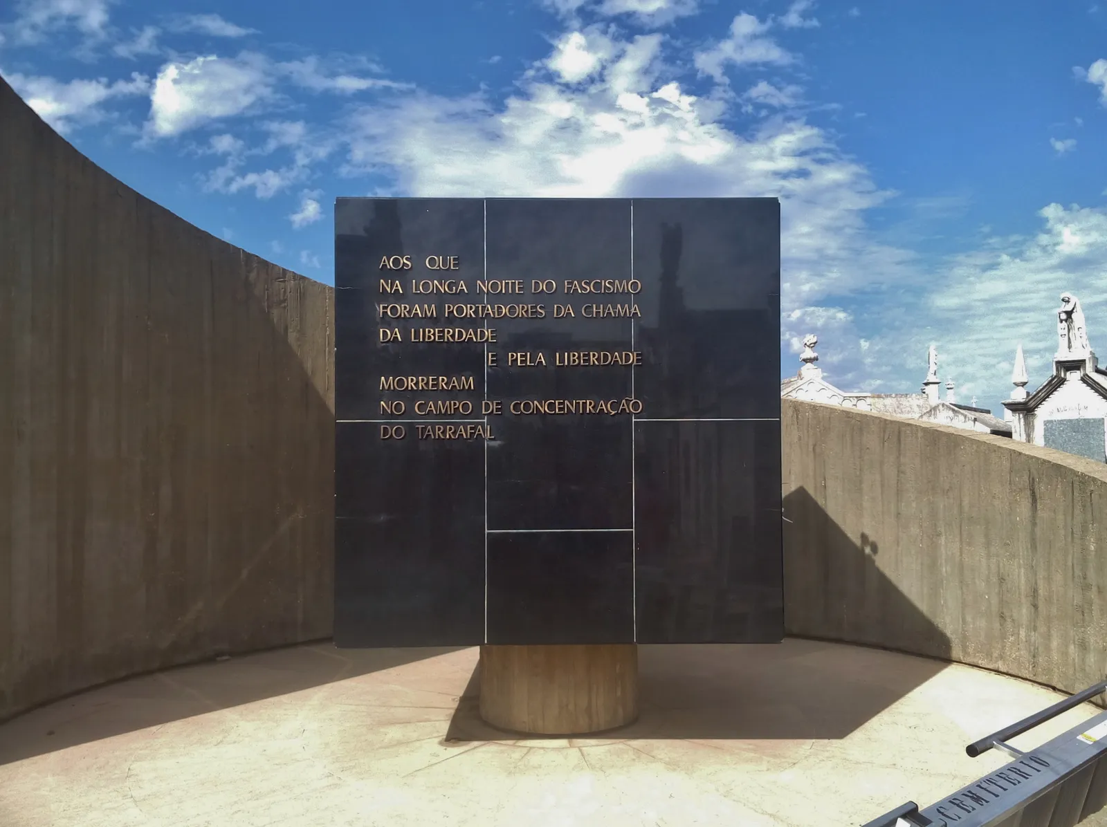 A large square slab of black stone with a gilt inscription in Portuguese stands on a round pedestal inside a curved concrete enclosure, under a blue sky with white clouds. The tops of cemetery chapels show over the wall on the right.