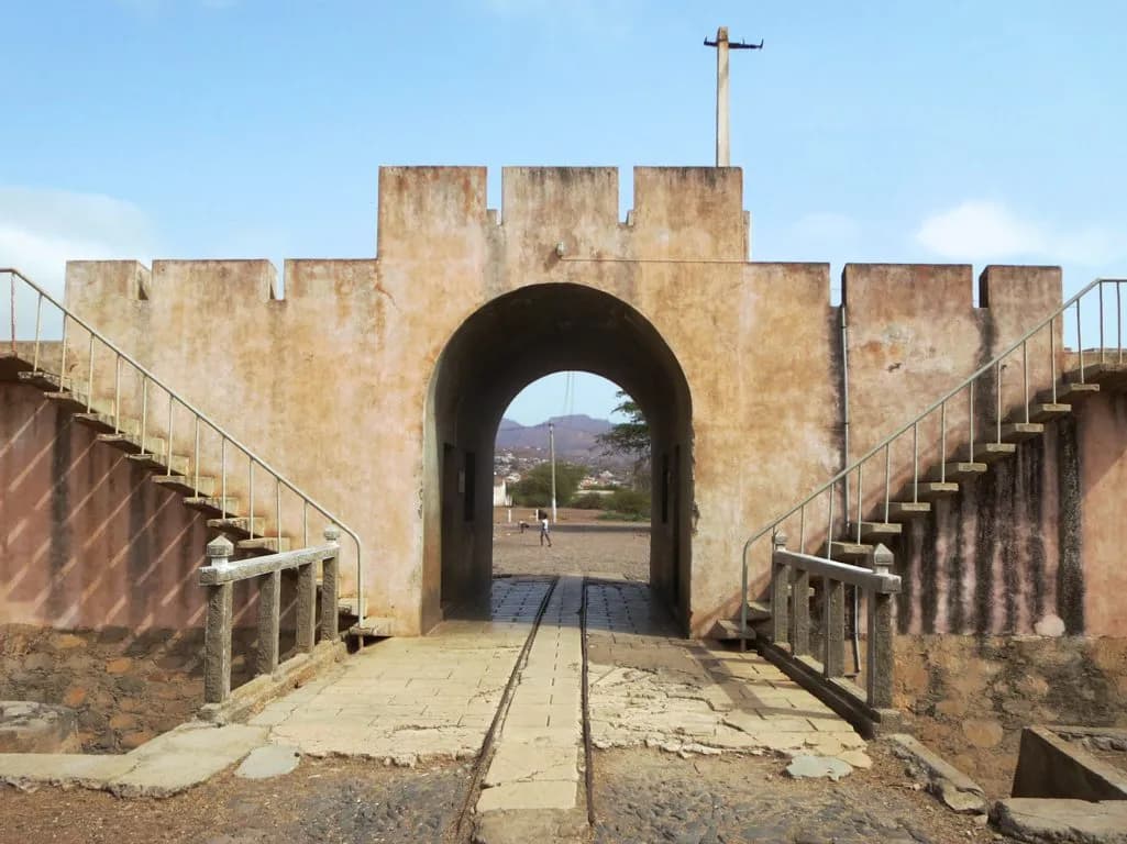 A weathered ochre gatehouse with crenellations and a round arch, reached by a short paved bridge with low stone railings. Flights of steps with metal handrails climb the wall on either side, a tall pole rises above, and hills show through the arch.