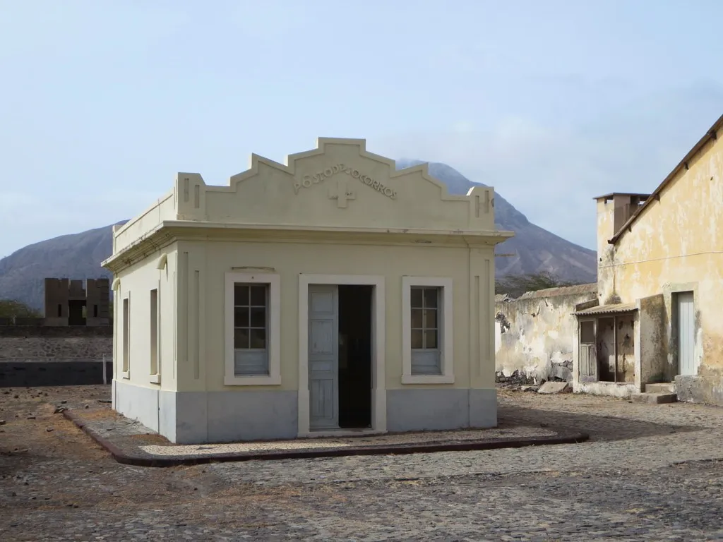 A small pale single-storey building with a stepped gable, an open door between two windows, and the words Posto de Socorros above a cross on the facade. It stands on cobbles beside a weathered ochre building, with a mountain behind.