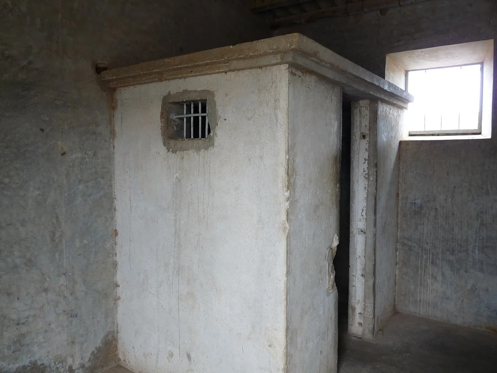 Inside a bare room with stained grey walls, a narrow whitewashed concrete box with a flat overhanging top and a small barred opening stands against the wall, its doorway at the side.