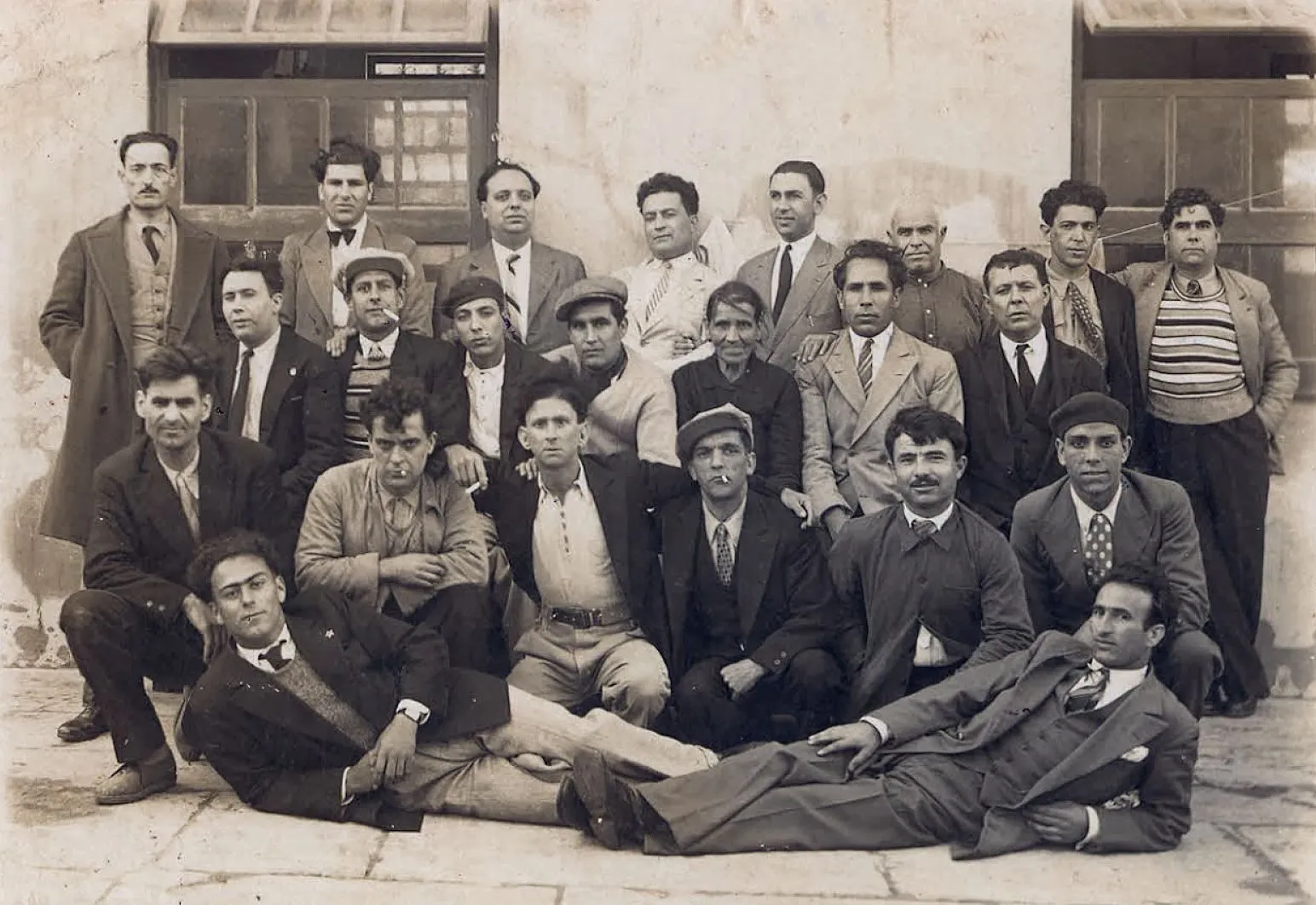 An old sepia group photograph of about two dozen people, most of them young men in suits, caps and striped jumpers, posing in rows against a whitewashed wall with two large windows. Two men lie on the ground in front.