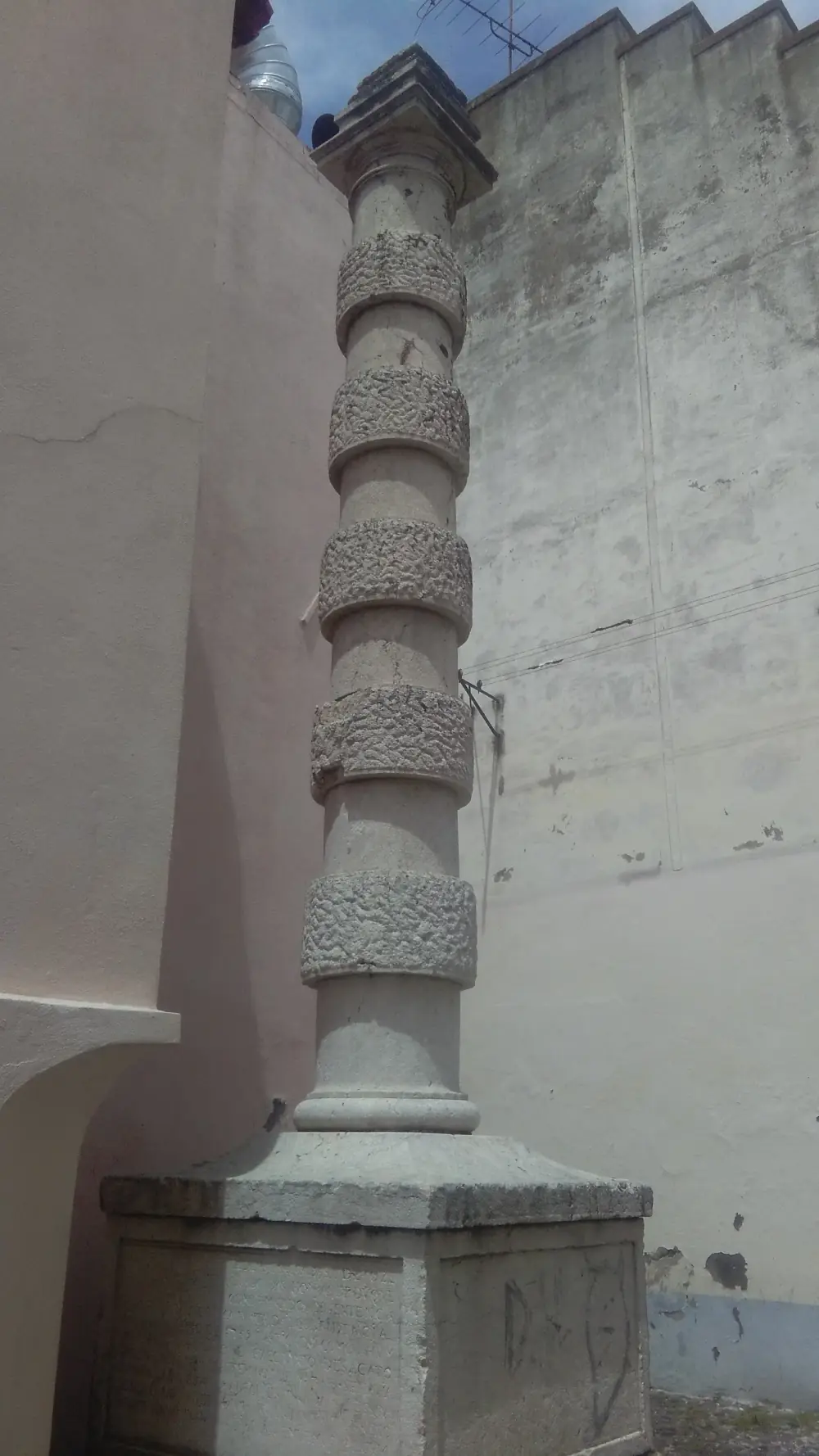 A slender stone column ringed by five bands of rough carved stone stands on a square plinth in a narrow space between plain rendered walls.