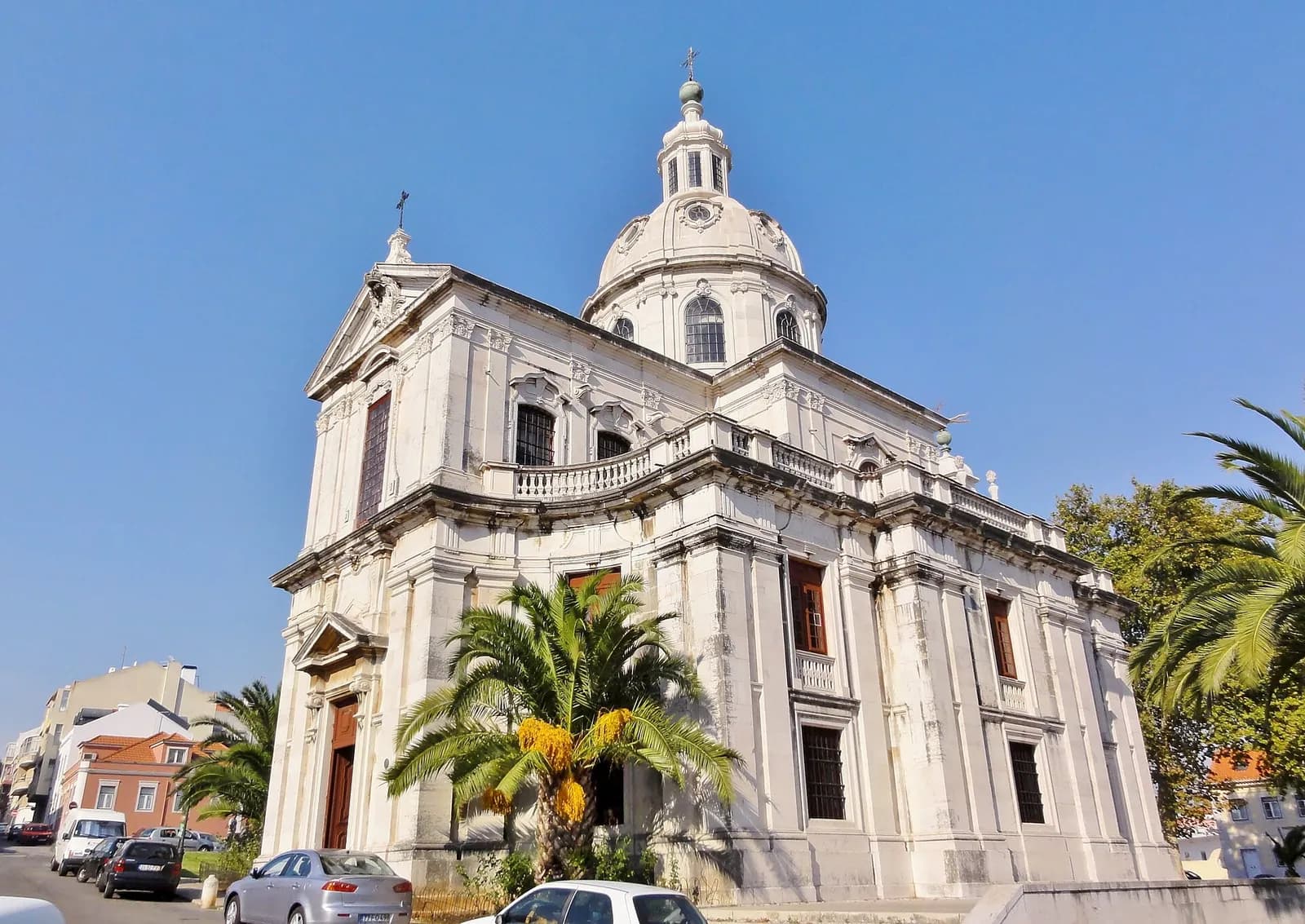 A white stone church with a classical facade, a balustrade along the roof and a dome topped by a small lantern and cross, seen against a clear blue sky with palm trees and parked cars in front.