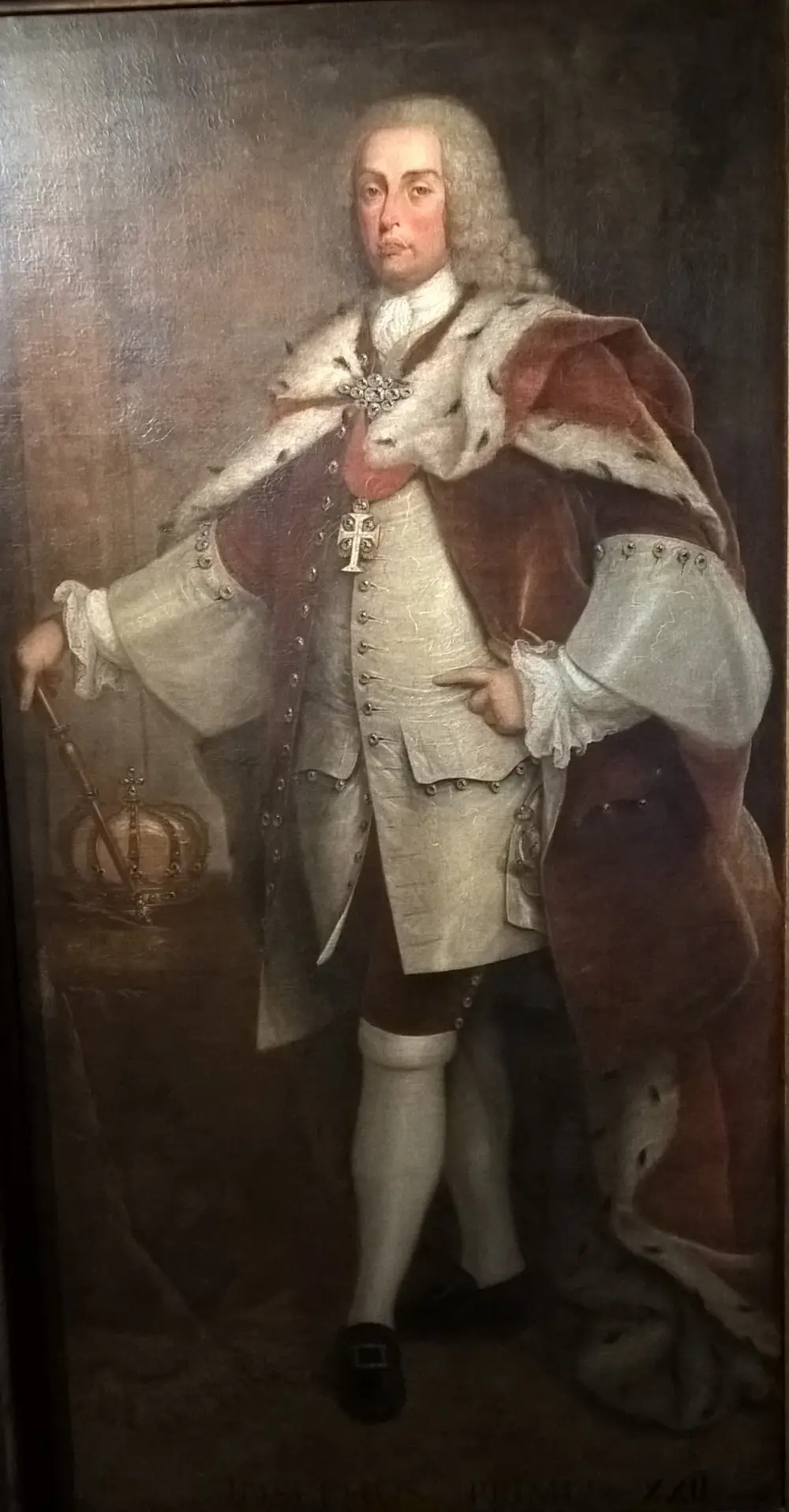 Full-length portrait of a heavy-set man in a powdered wig, a pale embroidered coat and breeches, with an ermine-lined red cloak over his shoulders. He rests one hand on a sceptre beside a crown on a cushion.
