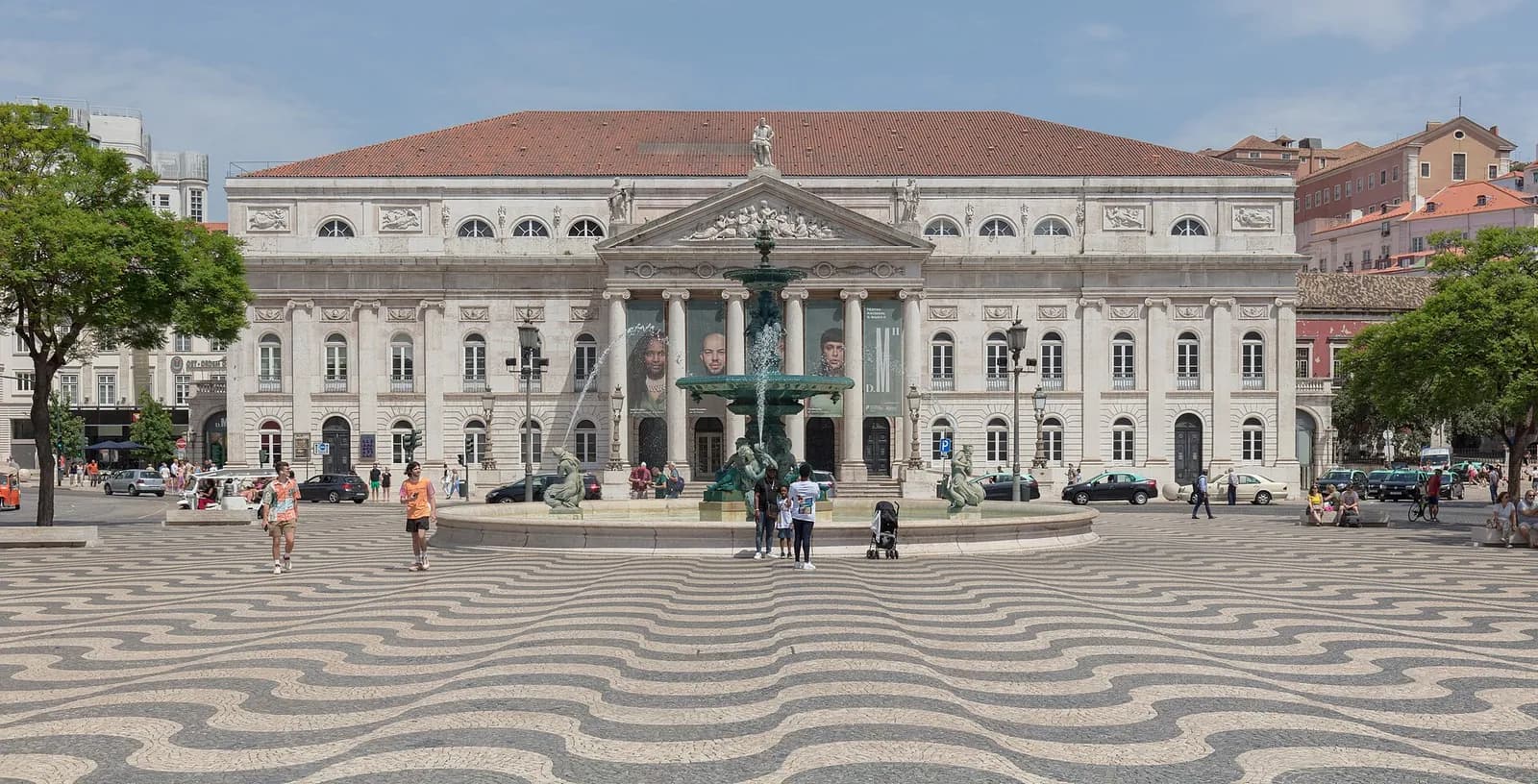 A long neoclassical building with a columned portico and triangular pediment, topped by a statue, closes the end of a square paved in black and white wave patterns. A fountain stands in front and people walk across the square in sunshine.
