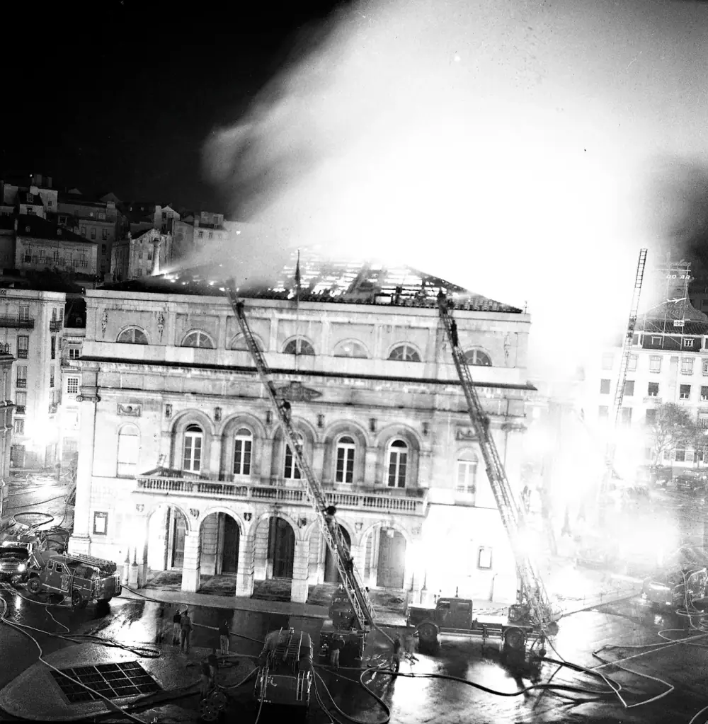 Night photograph of a large stone building with arched windows and an arcade at street level. Firefighters climb two tall fire-engine ladders that reach up to its roof, which is ablaze under a huge plume of lit smoke. Hoses snake across the wet square in front, where fire engines and a few figures stand.