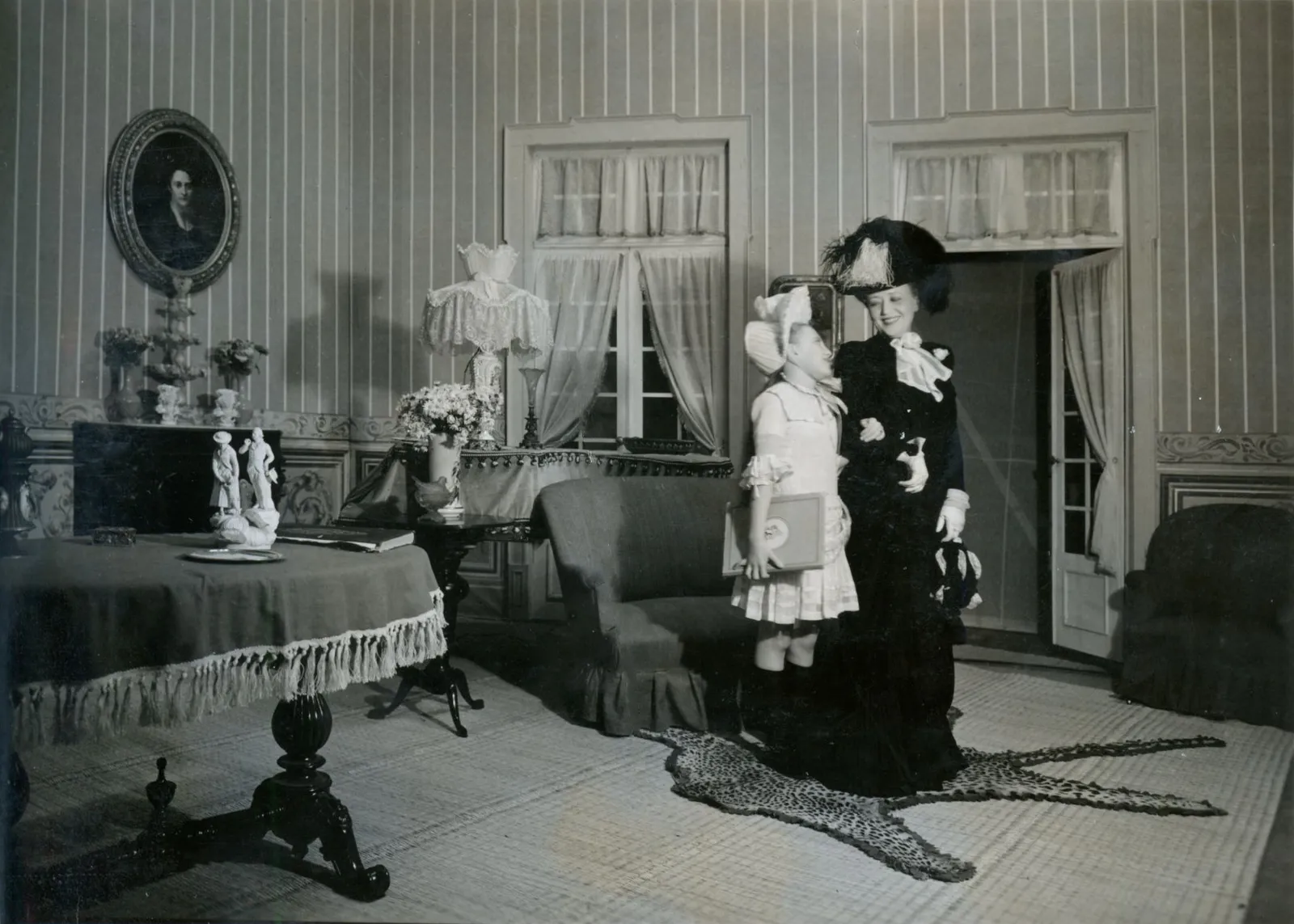A stage set of a nineteenth-century drawing room with striped wallpaper, a tasselled table and a leopard-skin rug. A woman in a long black dress and a large feathered hat stands beside a small girl in a white dress and bonnet who holds a book.