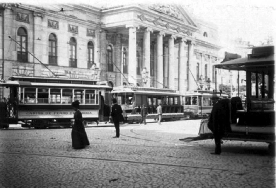 Black-and-white street scene: electric trams marked Carris de Ferro de Lisboa wait in front of a stone building with tall columns and arched windows. A woman in a long dark dress and a man in a suit stand on the cobbles in the foreground.