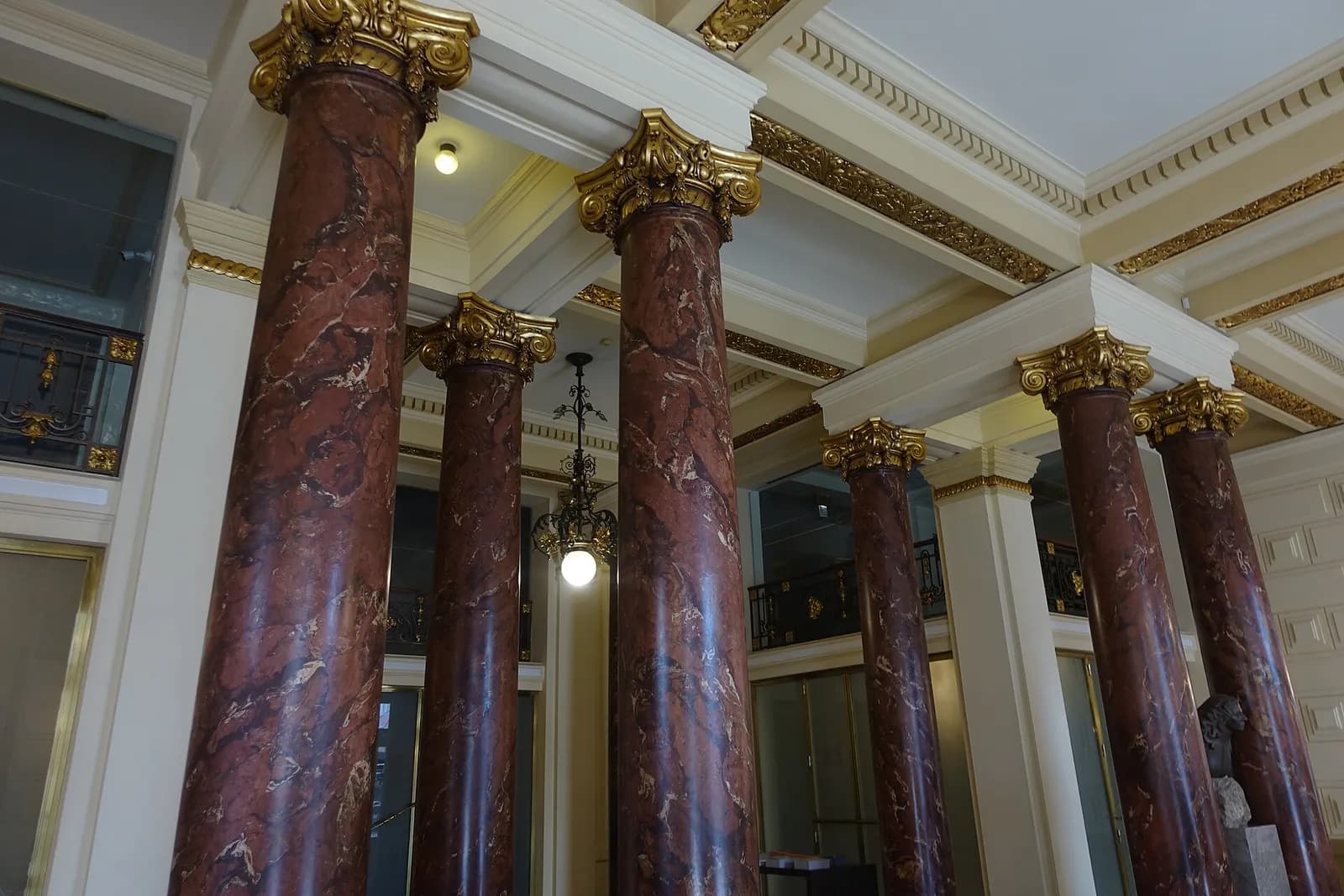 An interior with tall columns of polished red marbled stone topped by gilded capitals, between white walls and a coffered ceiling with gold mouldings; a lantern hangs between the columns.
