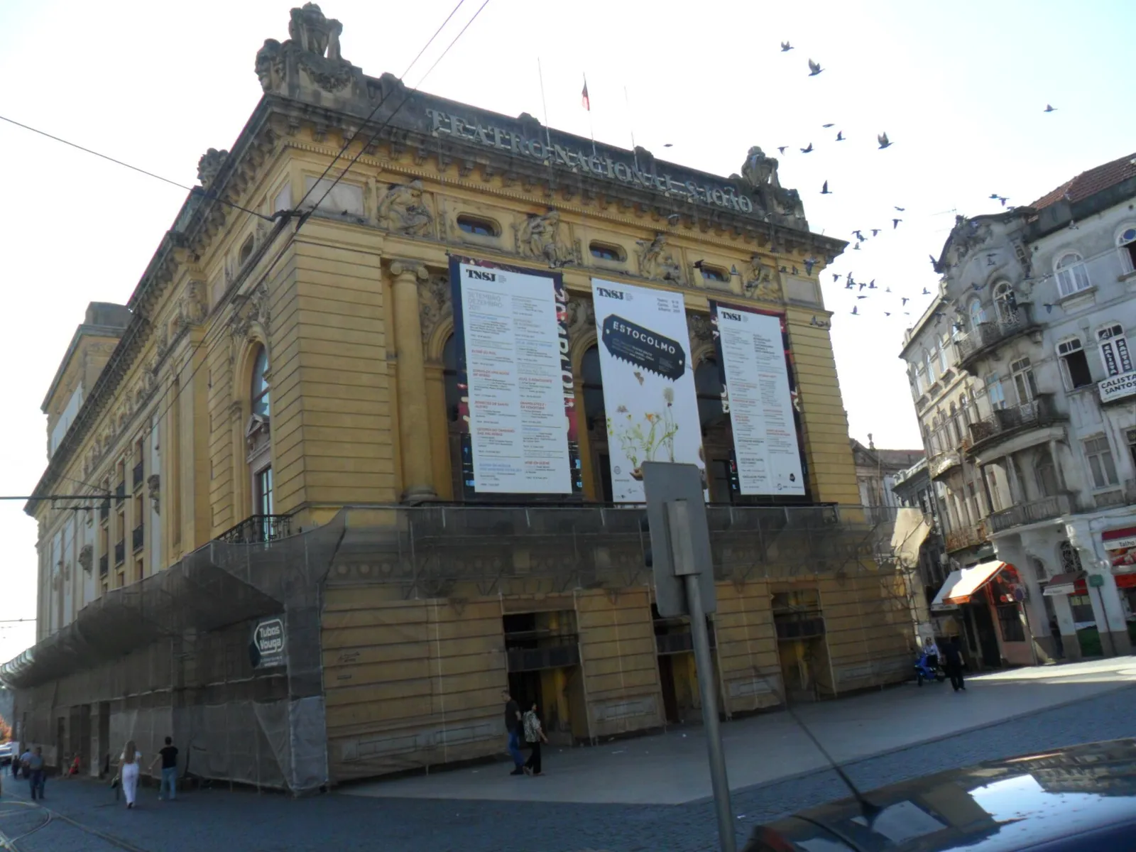 A yellow-painted theatre with a carved upper storey and three large programme banners on its front. The ground floor is hidden behind wooden hoarding and scaffolding wrapped in netting, and pigeons fly past on the right.